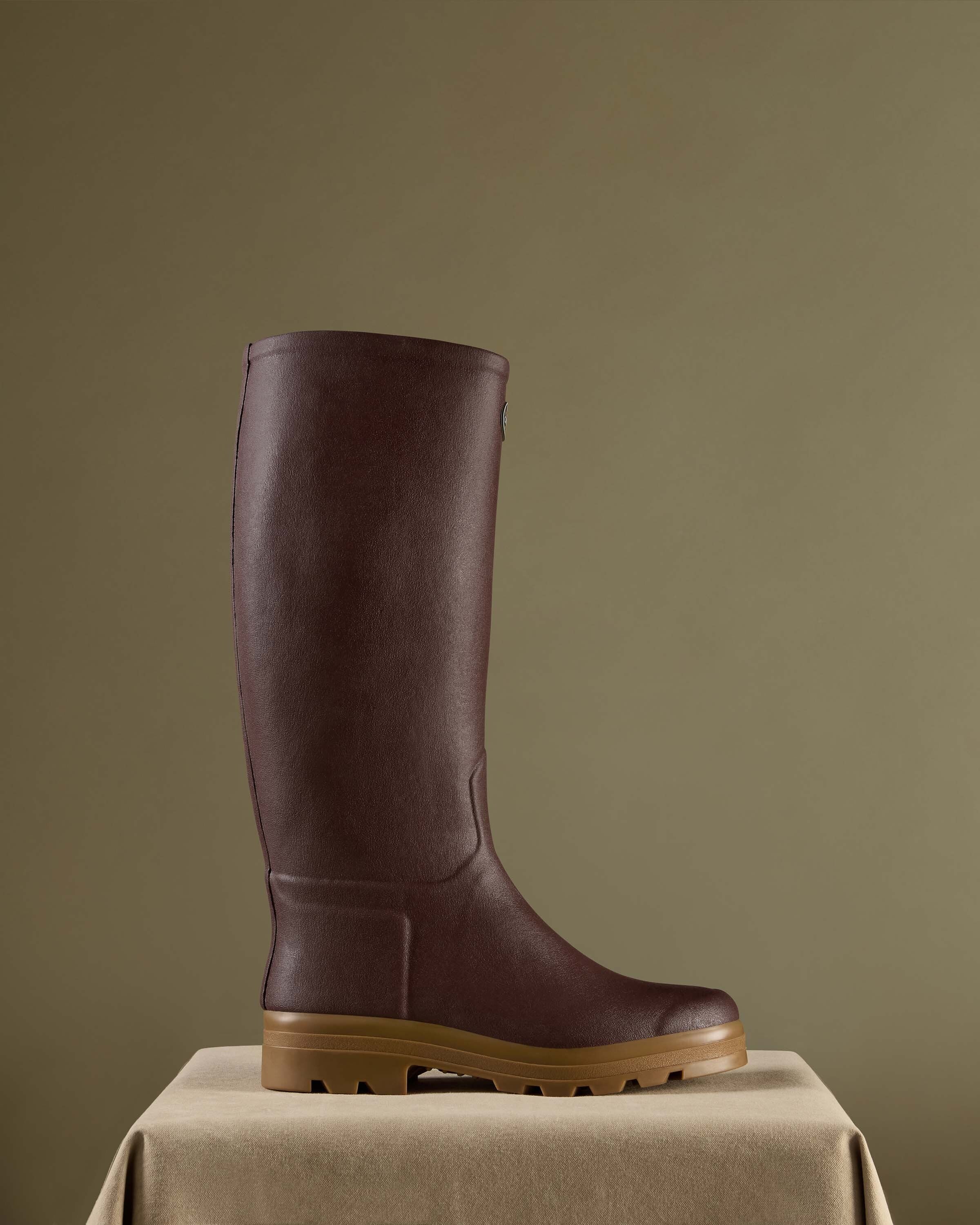 A single Men's Saint Hubert Leather Lined Wellington Boot in Brown displayed on a beige pedestal against an olive green background. The boot is photographed from the side to highlight its elegant, structured silhouette and premium finish.