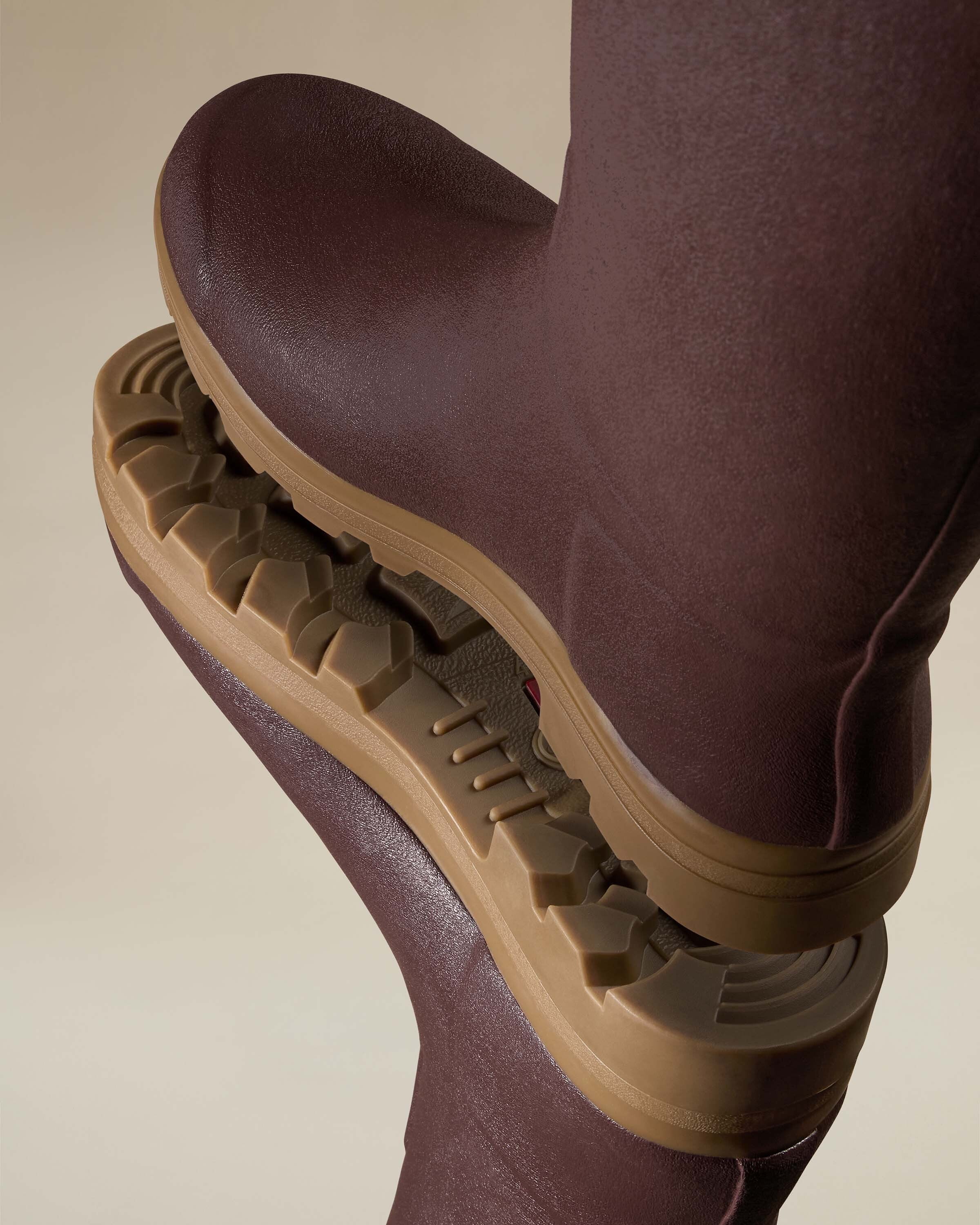 A dramatic close-up shot of the outsoles of two Men's Saint Hubert Leather Lined Wellington Boot in Brown pressed against each other to showcase the durable tread pattern. The tan sole provides contrast against the boot’s brown rubber upper.
