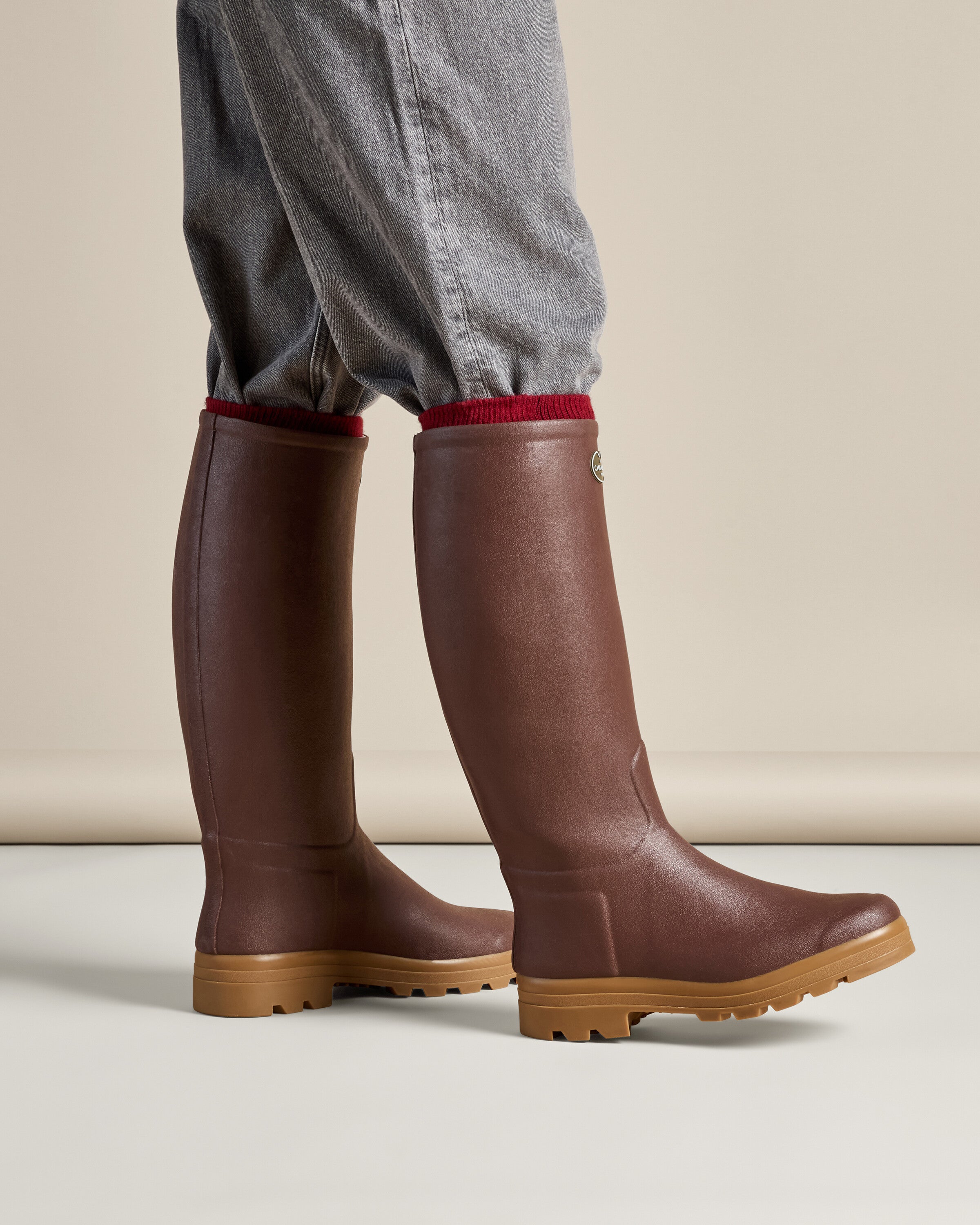 A lifestyle image of the Men's Saint Hubert Leather Lined Wellington Boot in Brown worn by a model with grey cuffed trousers tucked into the shafts. The setting is neutral, putting focus on the boots’ sleek structure and how they fit on the legs.