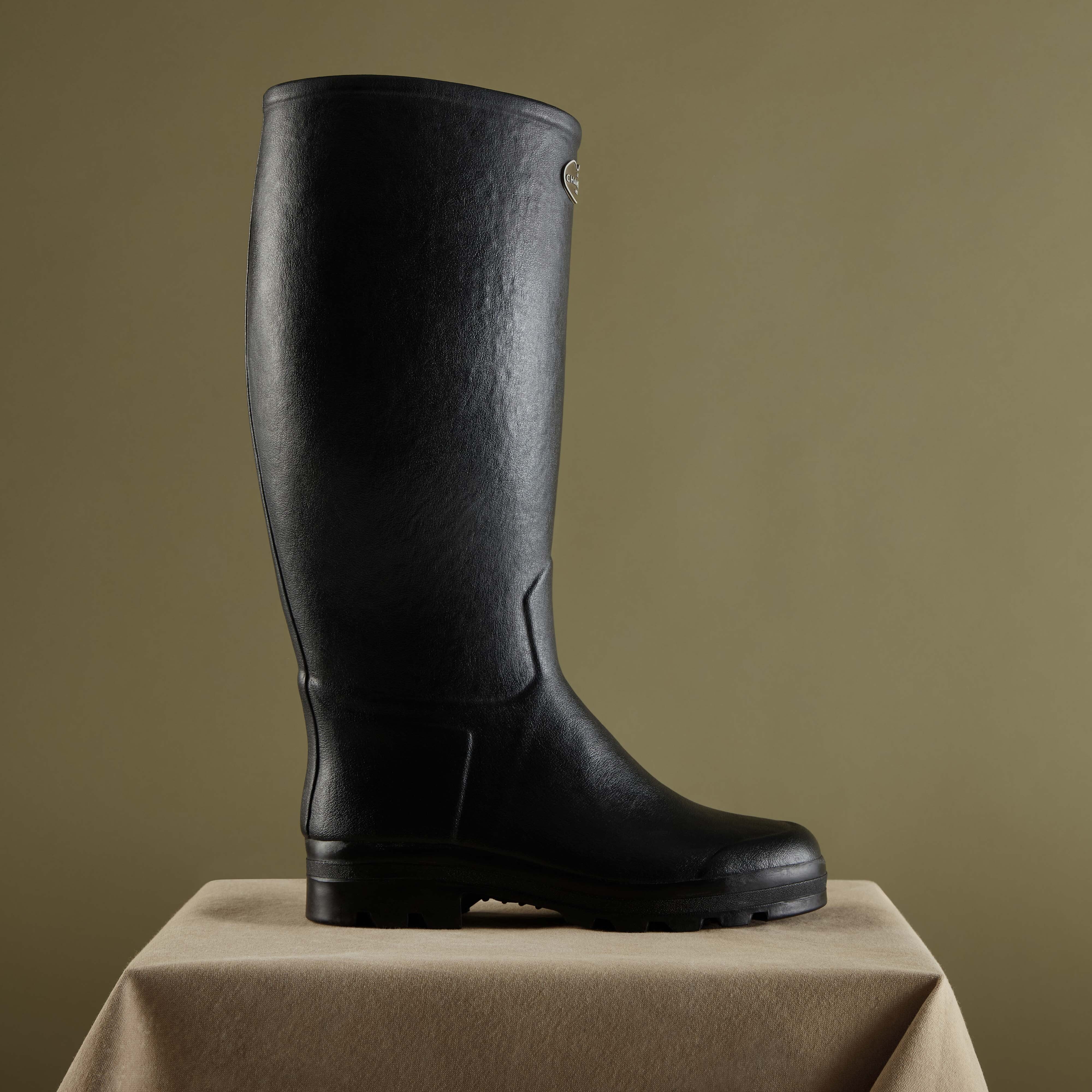 The same Men's Saint Hubert Leather Lined Wellington Boot in Black displayed on a neutral pedestal with an olive green background. The angled lighting highlights the natural texture of the rubber material and the soft matte finish.