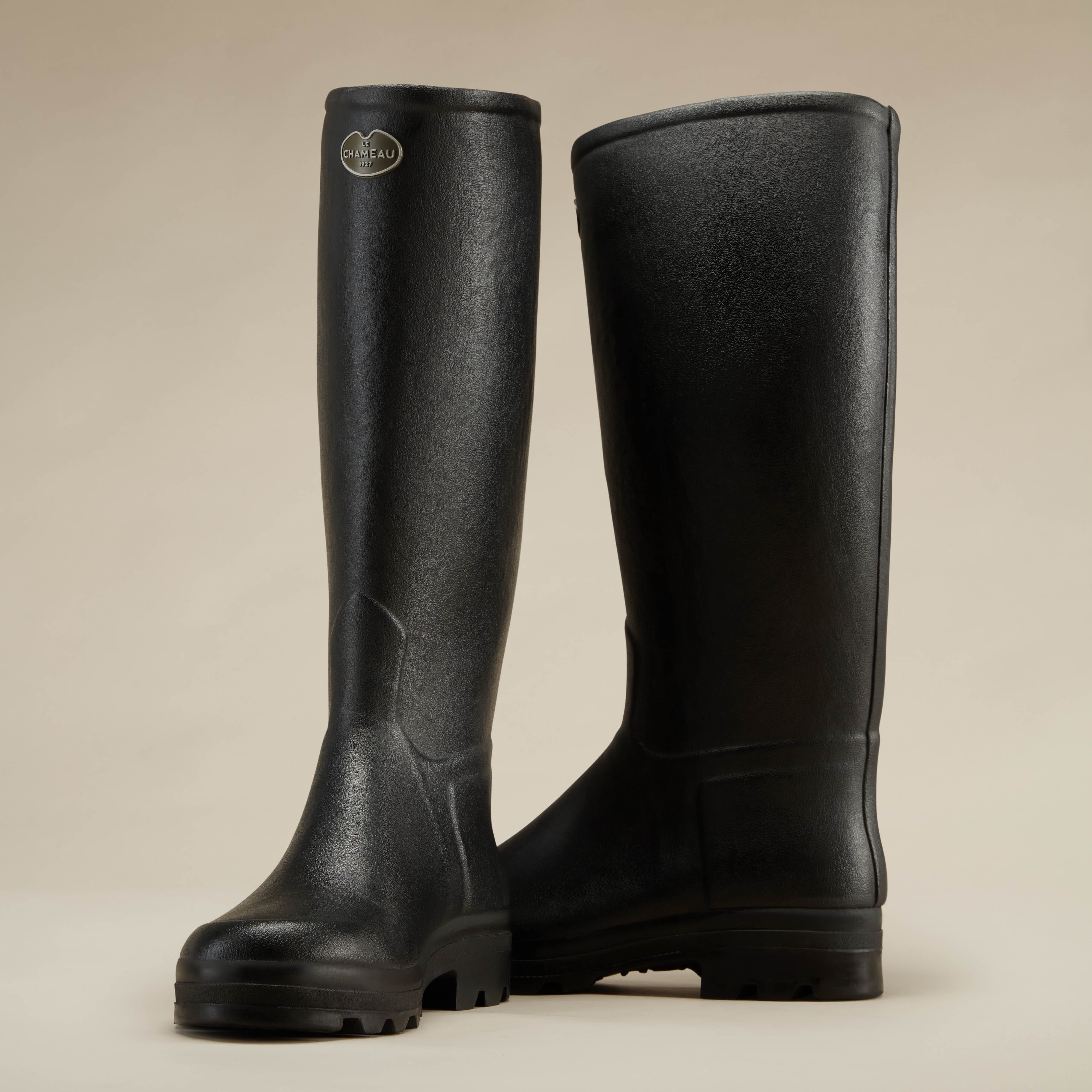 A pair of Men's Saint Hubert Leather Lined Wellington Boot in Black arranged to display both the front and back views on a beige background. The front boot shows the Le Chameau branding on the outer shaft, while the rear boot provides a clear view of the heel and back seam.