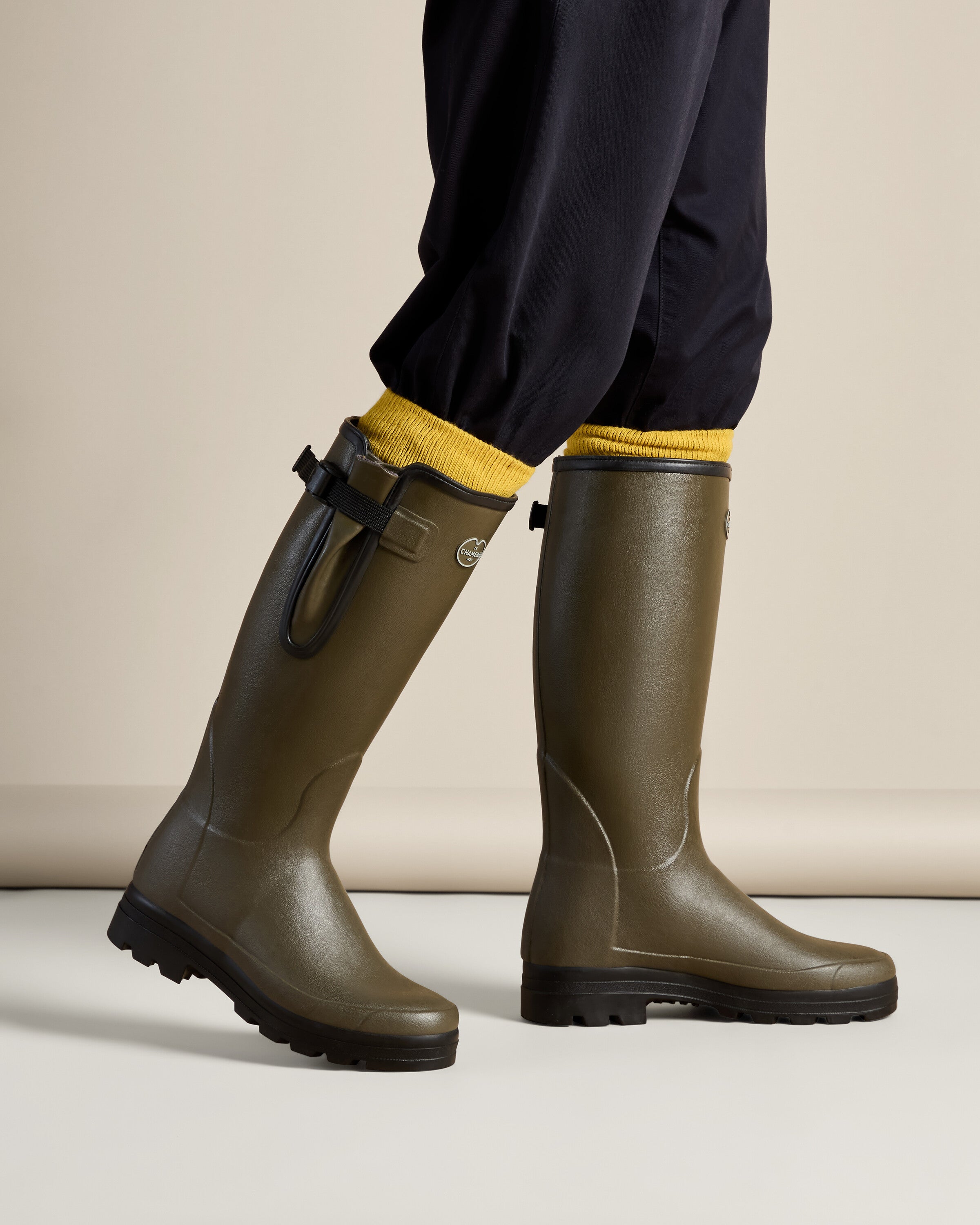 Side view of the Men's Vierzonord Neoprene Lined Wellington Boot in Chameau Green worn by a model. The black strap and gusset detail, combined with elasticated navy trousers tucked into the boots, underscore the functional elegance and snug fit ideal for all-weather outdoor activity.