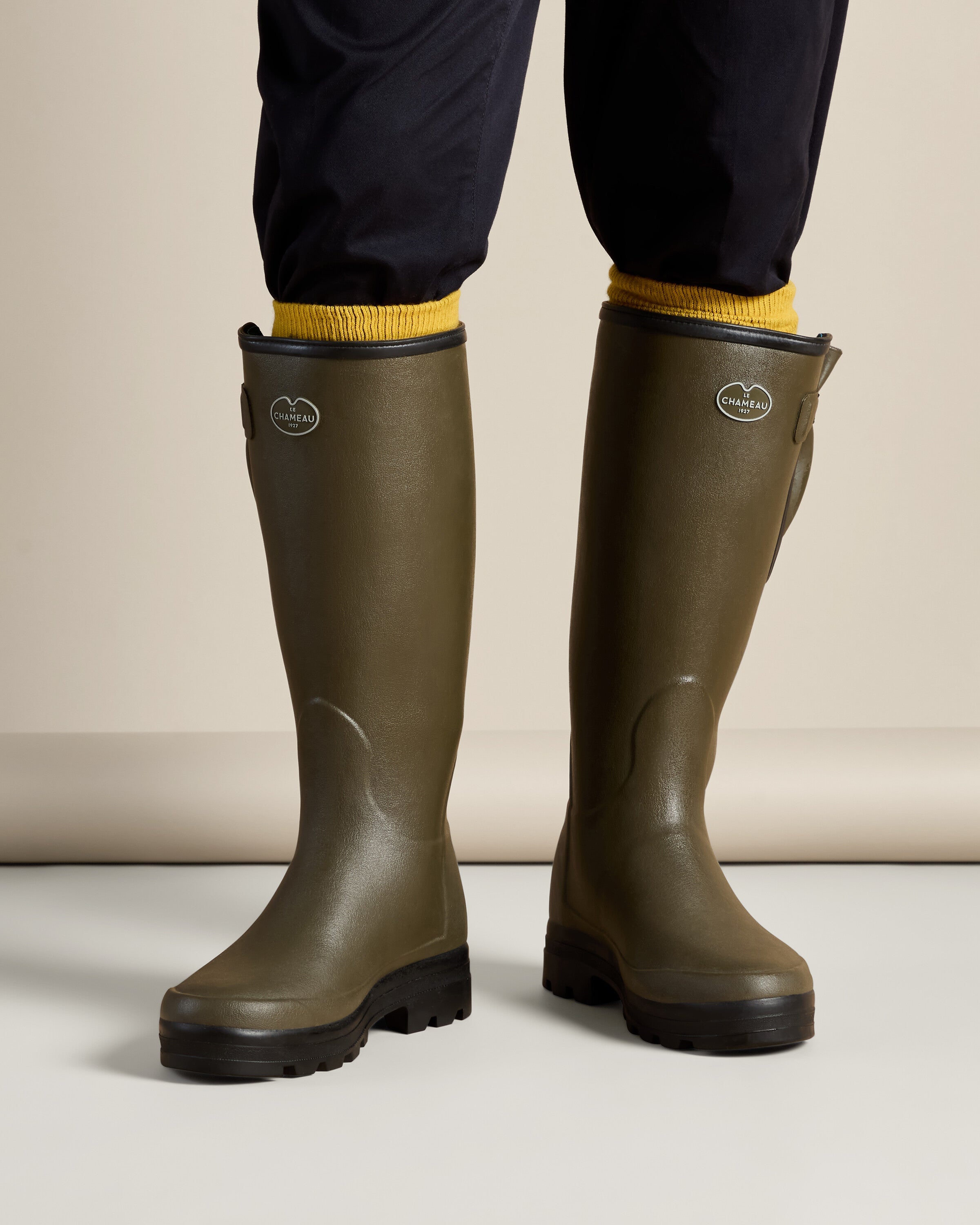 On-foot front view of a model wearing the Men's Vierzonord Neoprene Lined Wellington Boot in Chameau Green, paired with navy blue pants and yellow socks. The image highlights the comfortable fit and stylish contrast between functional outdoor footwear and casual wear.
