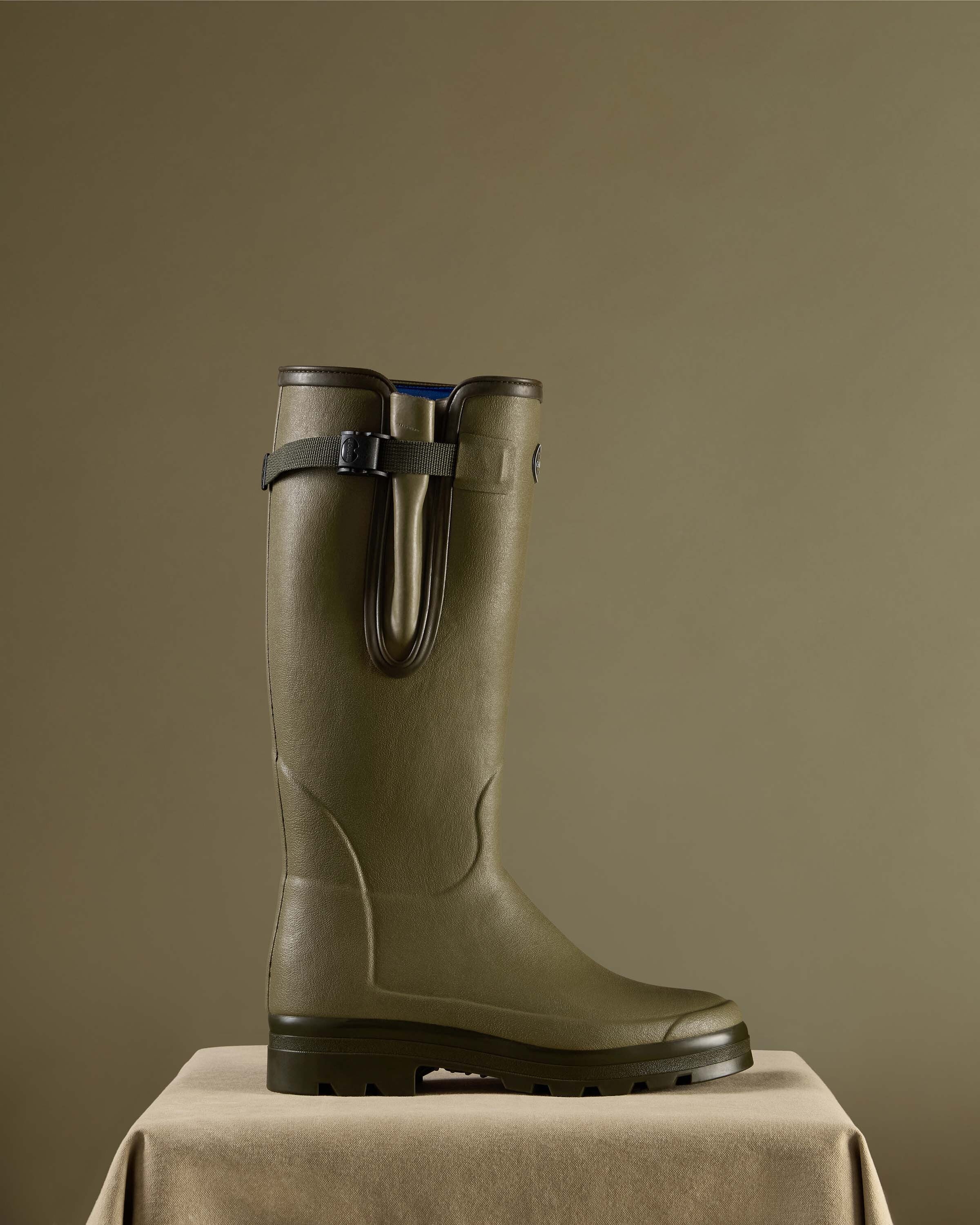 Single Men's Vierzonord Neoprene Lined Wellington Boot in Iconic Green displayed in full side profile on a soft beige pedestal with a moss green backdrop. The boot highlights its robust, handcrafted construction, rear gusset with strap, and deep-treaded outsole for all-terrain grip.