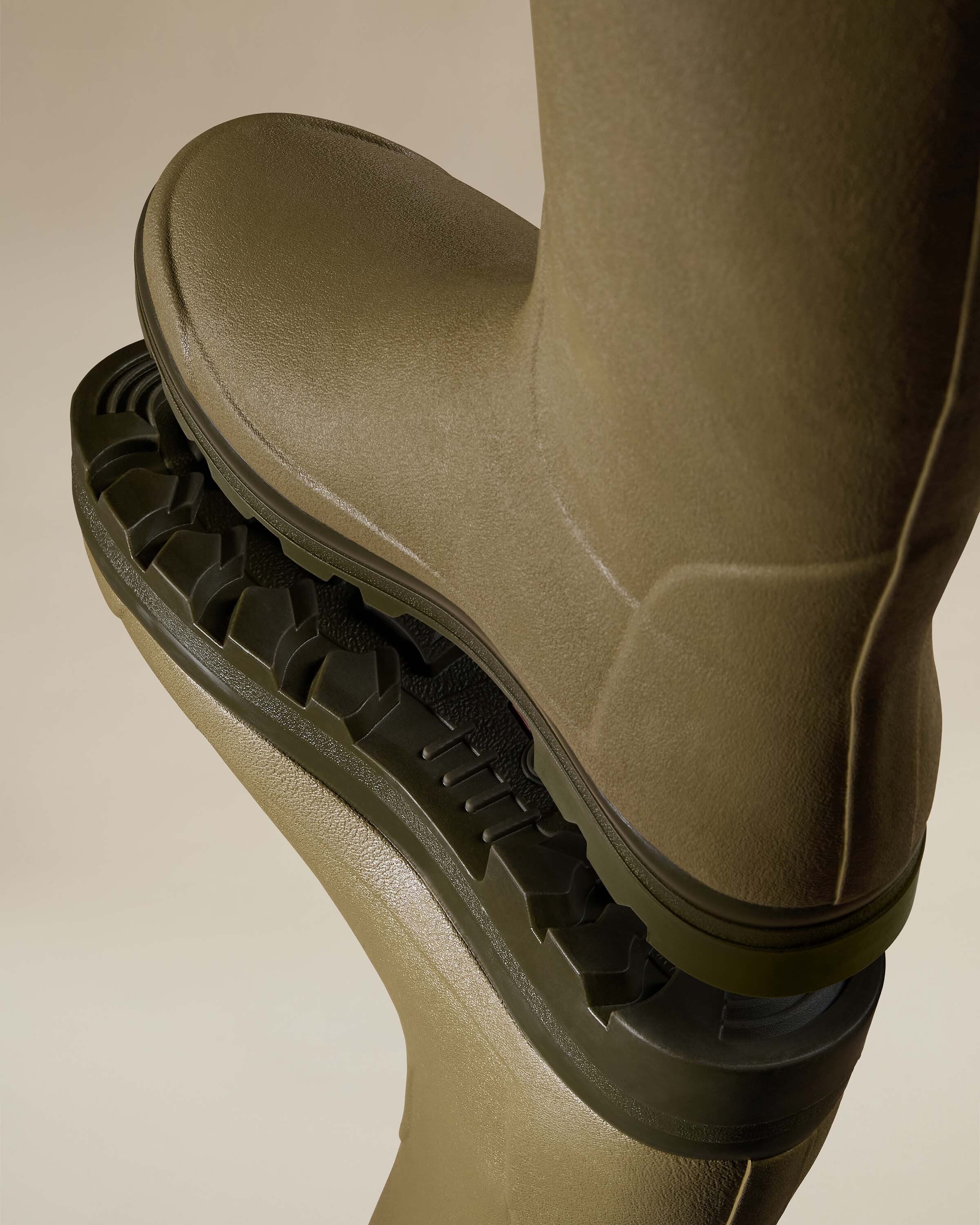 Close-up image showing the aggressive outsole tread of the Men's Vierzonord Neoprene Lined Wellington Boot in Iconic Green. One boot is stacked on top of another, sole upward, revealing the chunky multi-directional lugs and anti-slip design suitable for rugged terrain.