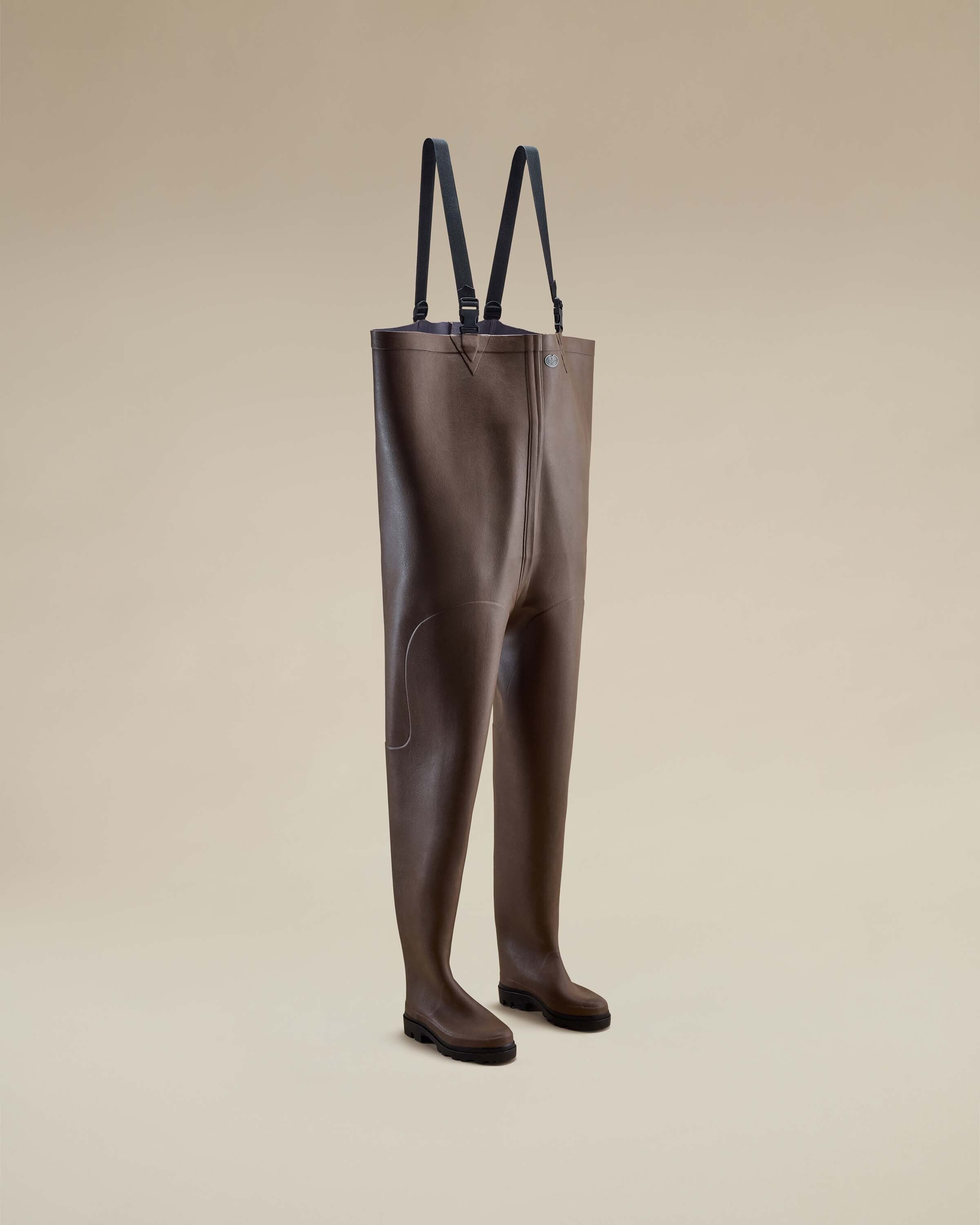 A three-quarter angle view of the Mer Renforcé Waders in Brown against a beige background, showing the front seam details and dual boot structure. The adjustable black straps are upright and clipped.