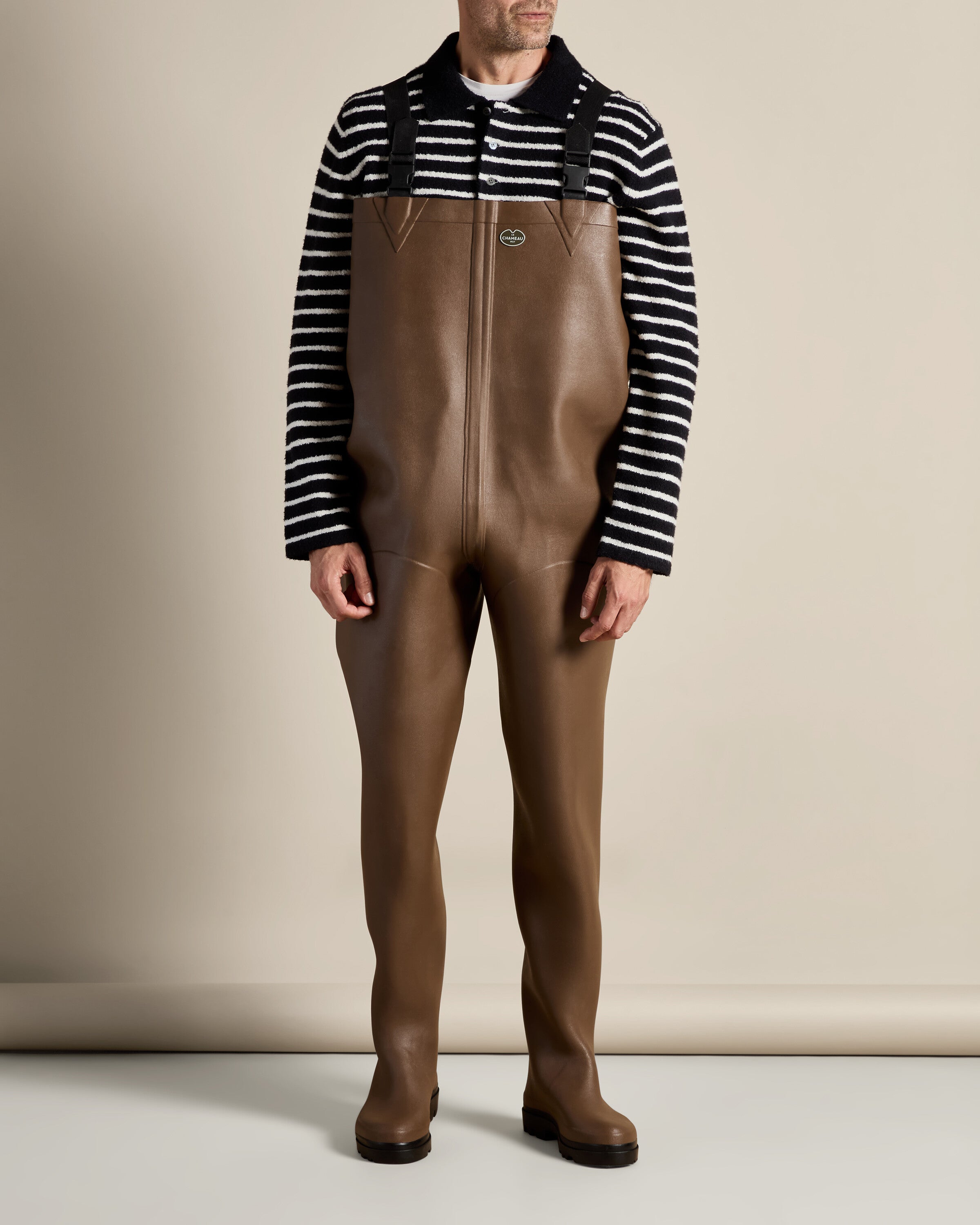 A male model wearing the Mer Renforcé Waders in Brown, standing front-facing against a neutral background. He is also wearing a black and white striped sweater, and the waders reach his chest.