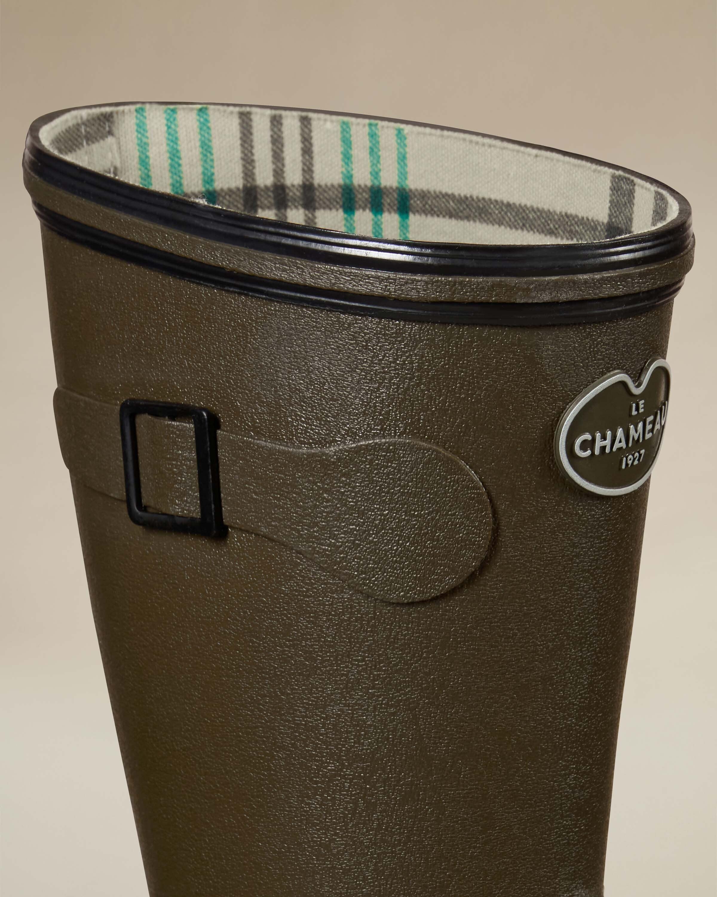 Close-up of the upper section of the Women's Giverny Jersey Lined Bottillon in Chameau Green, emphasizing the rubber texture, logo badge, and buckle strap. The jersey lining with teal and gray stripes is clearly visible inside the boot shaft.