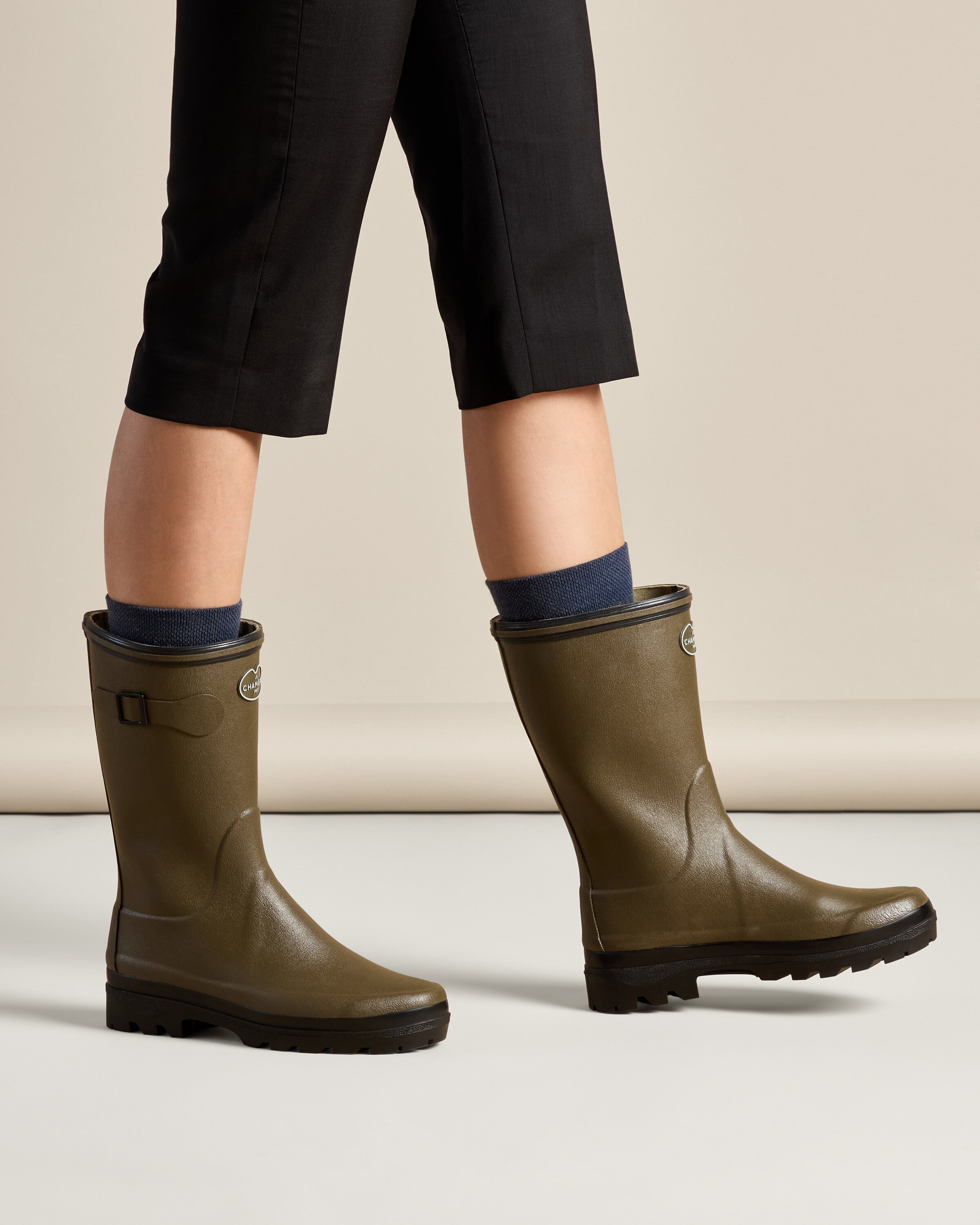 Side view of a model walking in the Women's Giverny Jersey Lined Bottillon in Chameau Green, paired with black cropped pants and navy socks. Both boots are visible in motion, demonstrating their comfort and sleek silhouette.