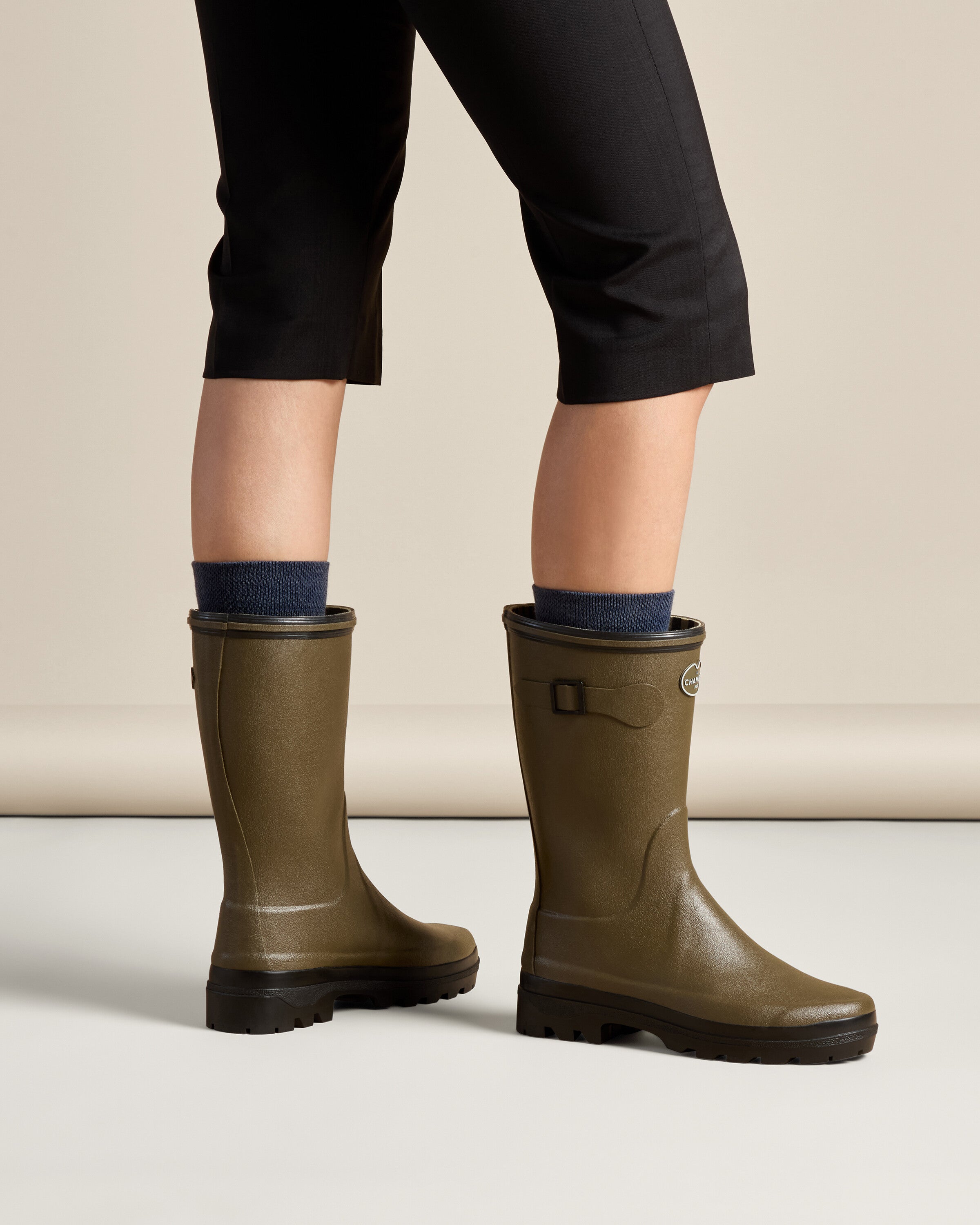 Rear view of a model wearing the Women's Giverny Jersey Lined Bottillon in Chameau Green with navy socks and cropped black pants. The boots' fit and height on the legs are showcased, as well as the simple and sturdy design.