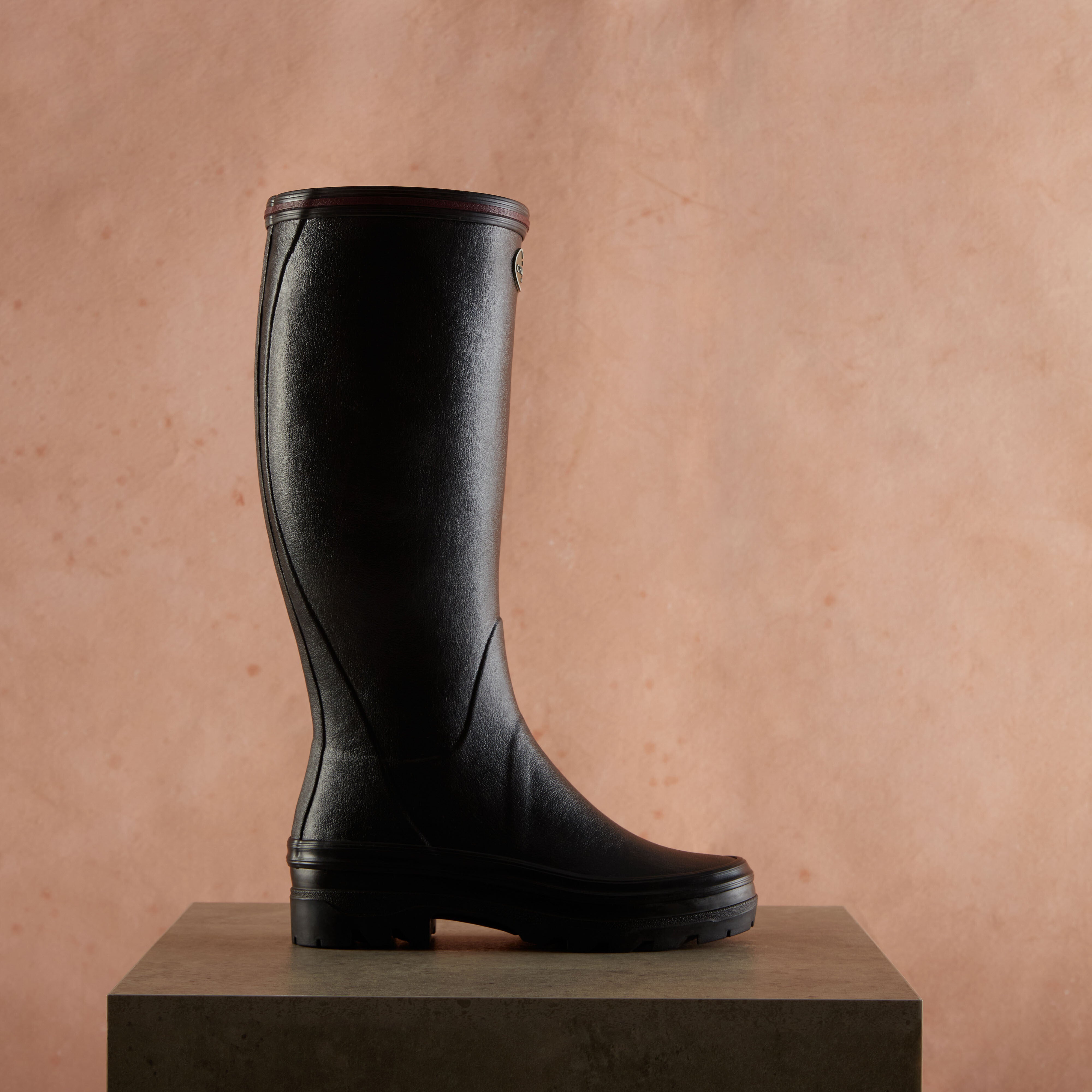 Single black Women's Giverny Jersey Lined Wellington Boot in Black shown in full profile, placed on a dark grey pedestal with a warm pink textured wall background. The boot appears sleek and elegant, designed for both durability and style.
