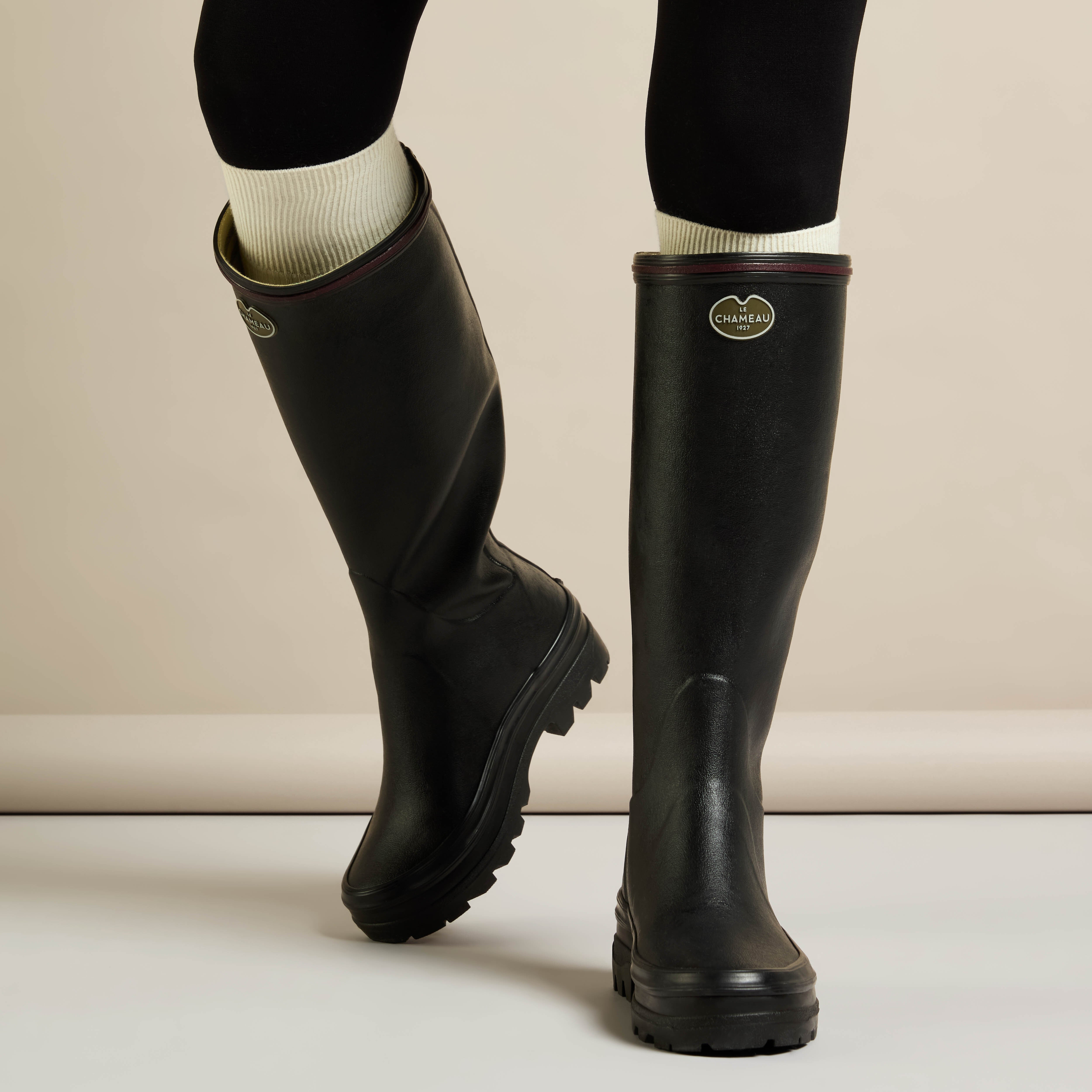 Lifestyle shot of a model wearing the Women's Giverny Jersey Lined Wellington Boot in Black, paired with cream ribbed socks and black leggings. The model is mid-step, showcasing the boots’ flexible yet structured fit and the brand badge clearly displayed on both boots.