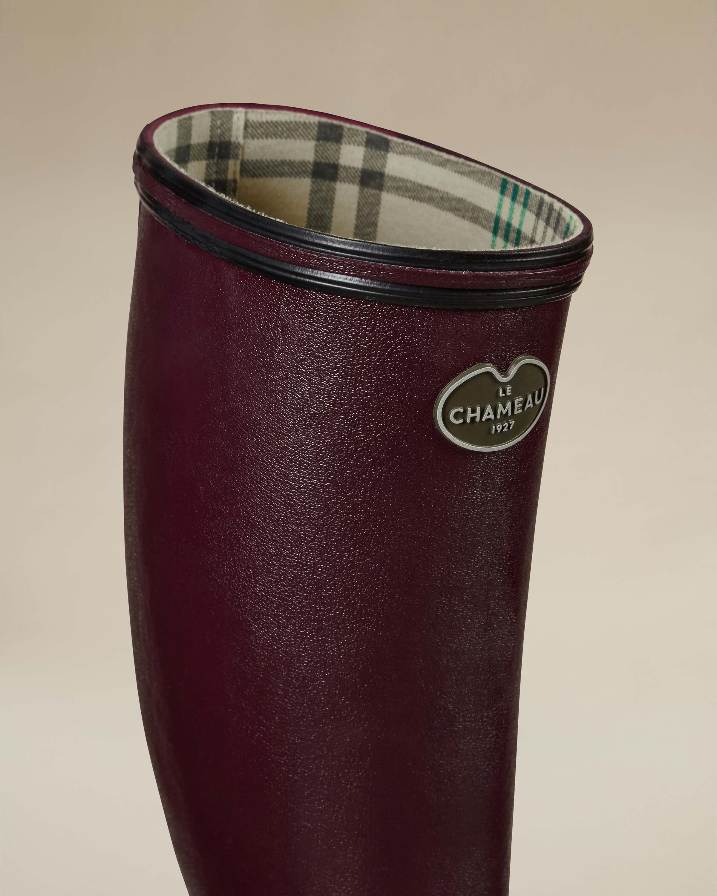Close-up of the upper shaft of a Women's Giverny Jersey Lined Wellington Boot in Cherry Red, focusing on the brand’s logo badge and the boot's opening. The cream-colored jersey lining with green and grey plaid stripes is visible inside the boot.