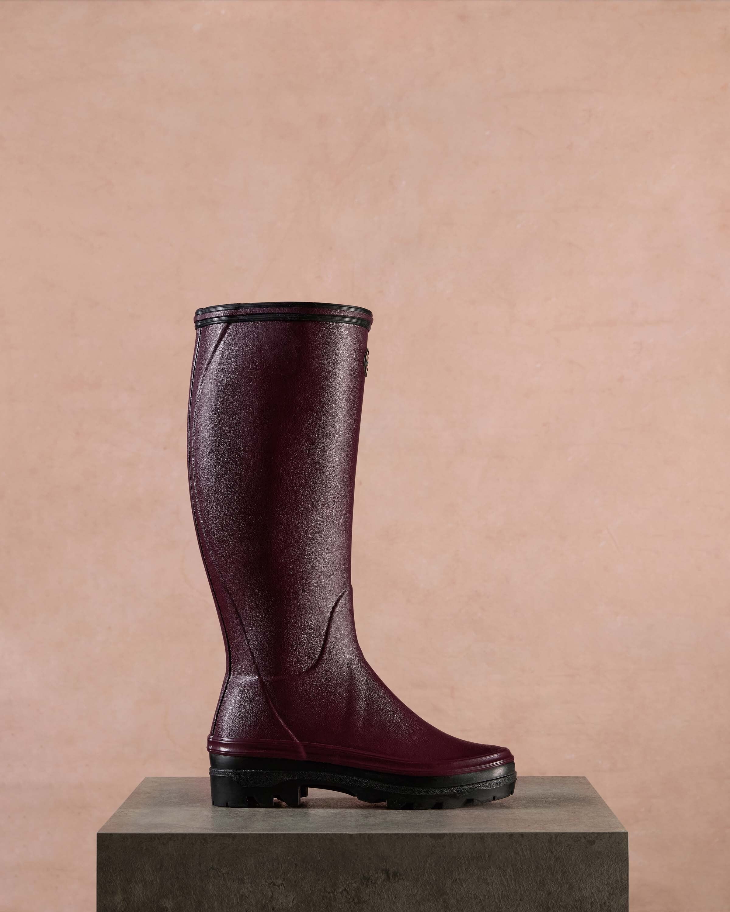 Single Women's Giverny Jersey Lined Wellington Boot in Cherry Red in profile placed on a dark stone pedestal against a soft pink textured background. The boot’s form is upright, highlighting its height, smooth curve, and robust treaded sole.