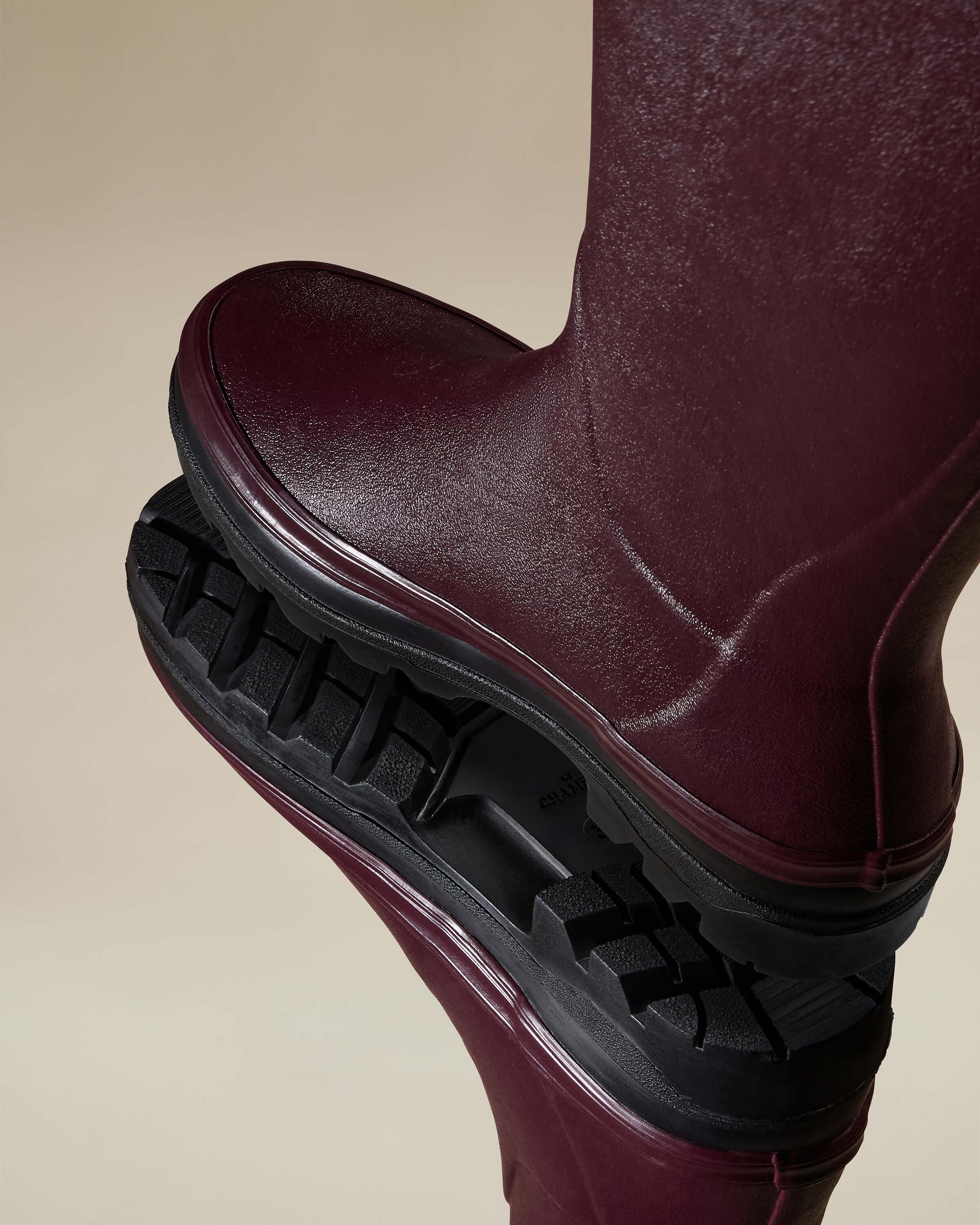 Dynamic close-up showing the sole of one Women's Giverny Jersey Lined Wellington Boot in Cherry Red stacked on another. The deep black tread grooves are highlighted in the composition, emphasizing the rugged and durable outsole.