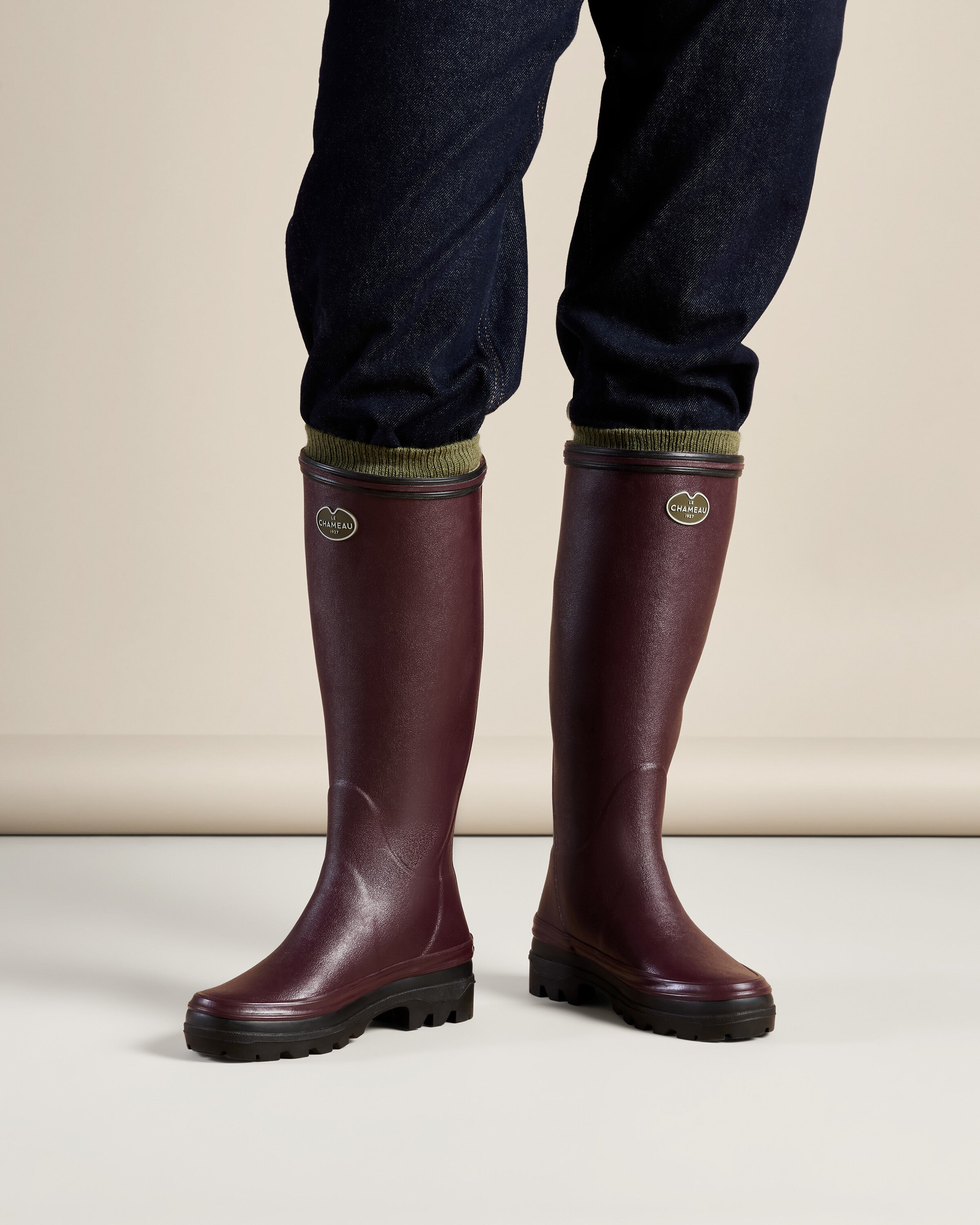 Model wearing a pair of Women's Giverny Jersey Lined Wellington Boot in Cherry Red with dark denim pants tucked into the boot shafts. The image focuses on the lower legs and feet, showcasing the boots in a lifestyle context, emphasizing fit and styling versatility.