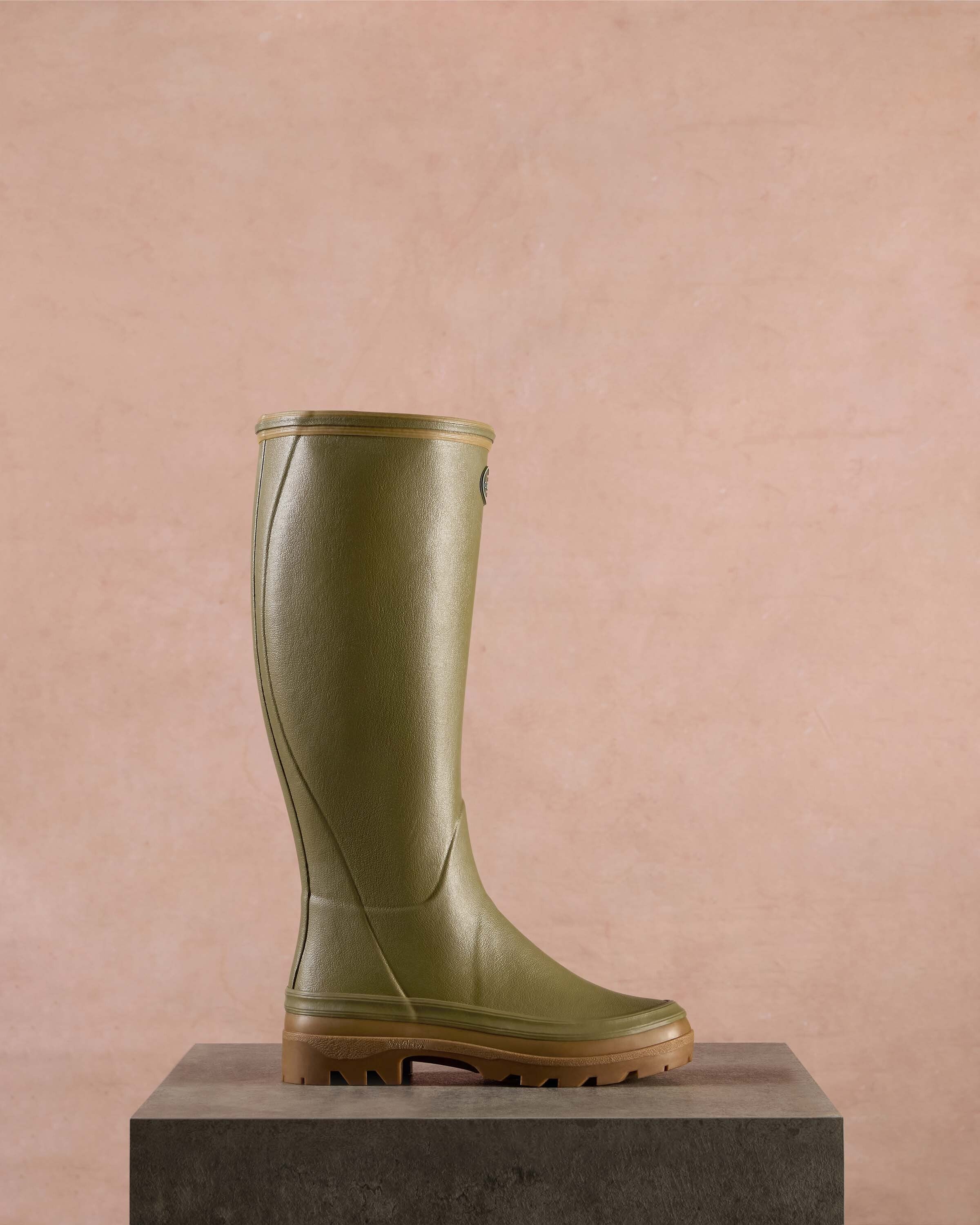 The Women's Giverny Jersey Lined Wellington Boot in Iconic Green is presented in a classic side profile while standing on a textured stone pedestal. The backdrop is a warm blush-toned wall, giving the product a premium, editorial feel. The shape and silhouette of the boot are emphasised with soft shadows.
