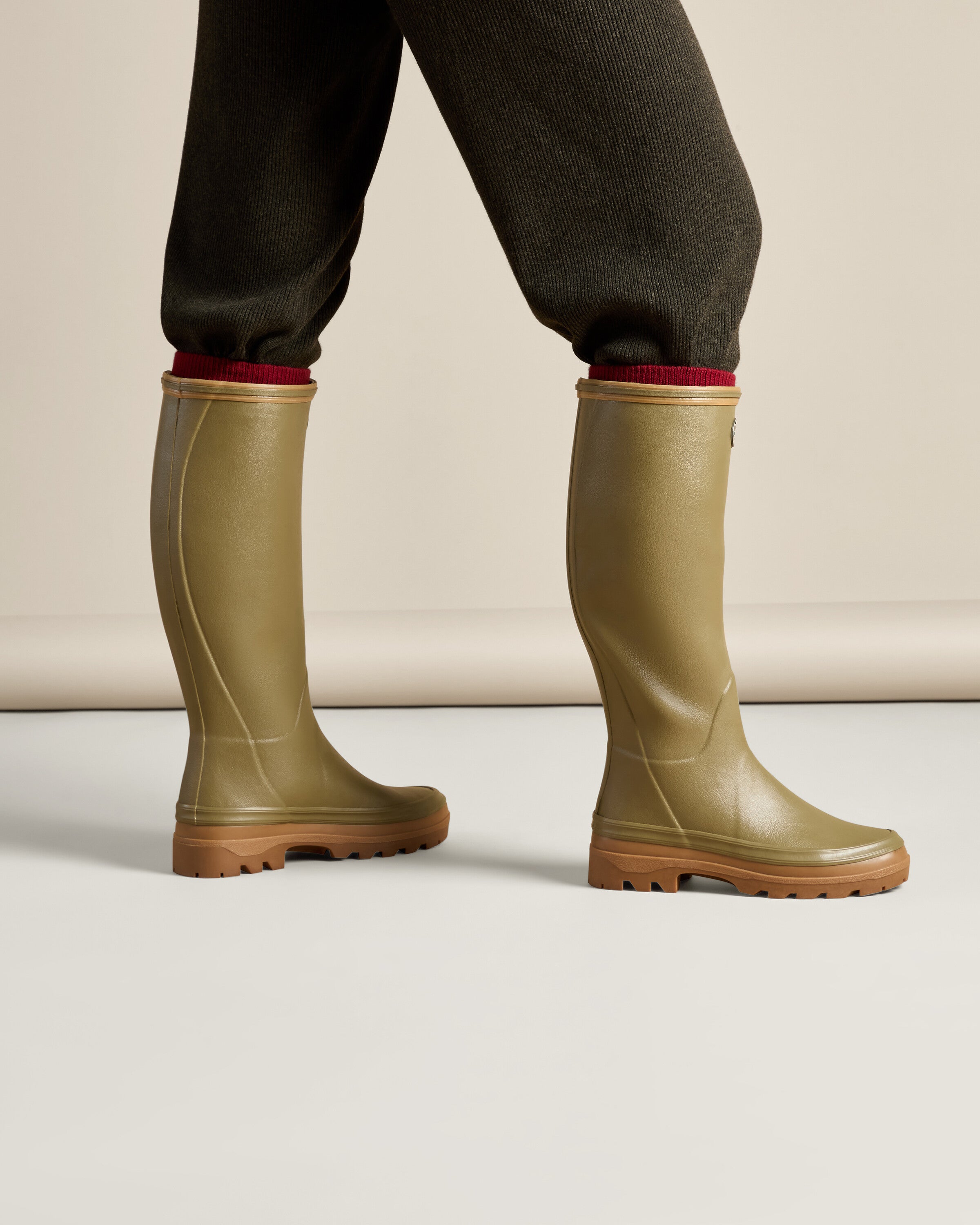 A model wears the Women's Giverny Jersey Lined Wellington Boot in Iconic Green with ribbed brown pants tucked into the shafts. The model is shown in a walking motion from the side, demonstrating the flexibility and practical height of the boots. The red sock rim peeks out for a touch of contrast.