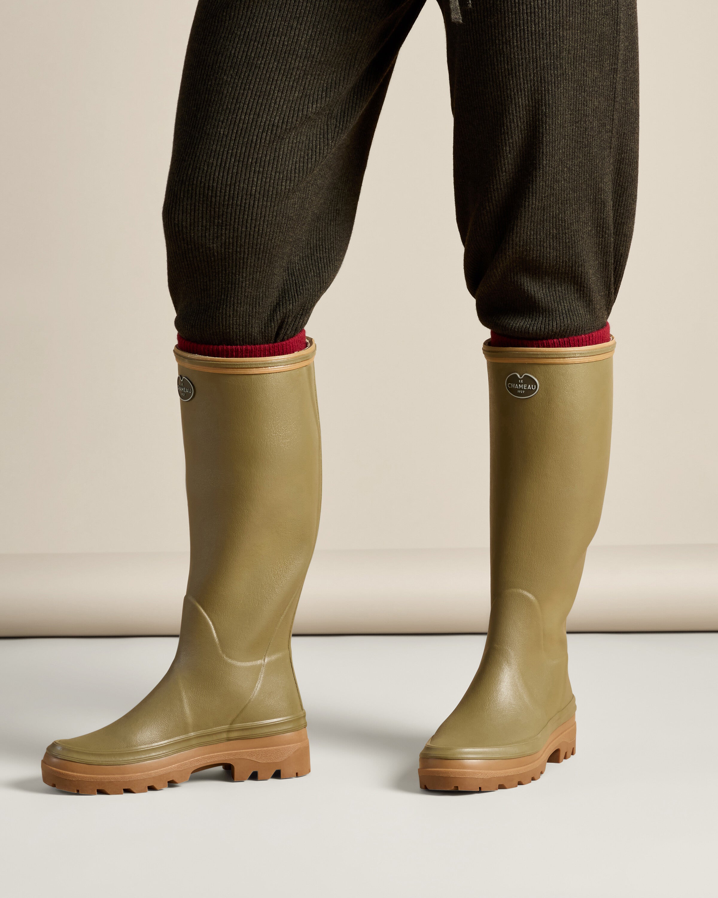 The model stands facing forward wearing the Women's Giverny Jersey Lined Wellington Boot in Iconic Green, allowing for a clear view of the boot's front profile. The soft gleam of the rubber material, the structured silhouette, and the stylish look of the boots are clearly presented in a lifestyle context.