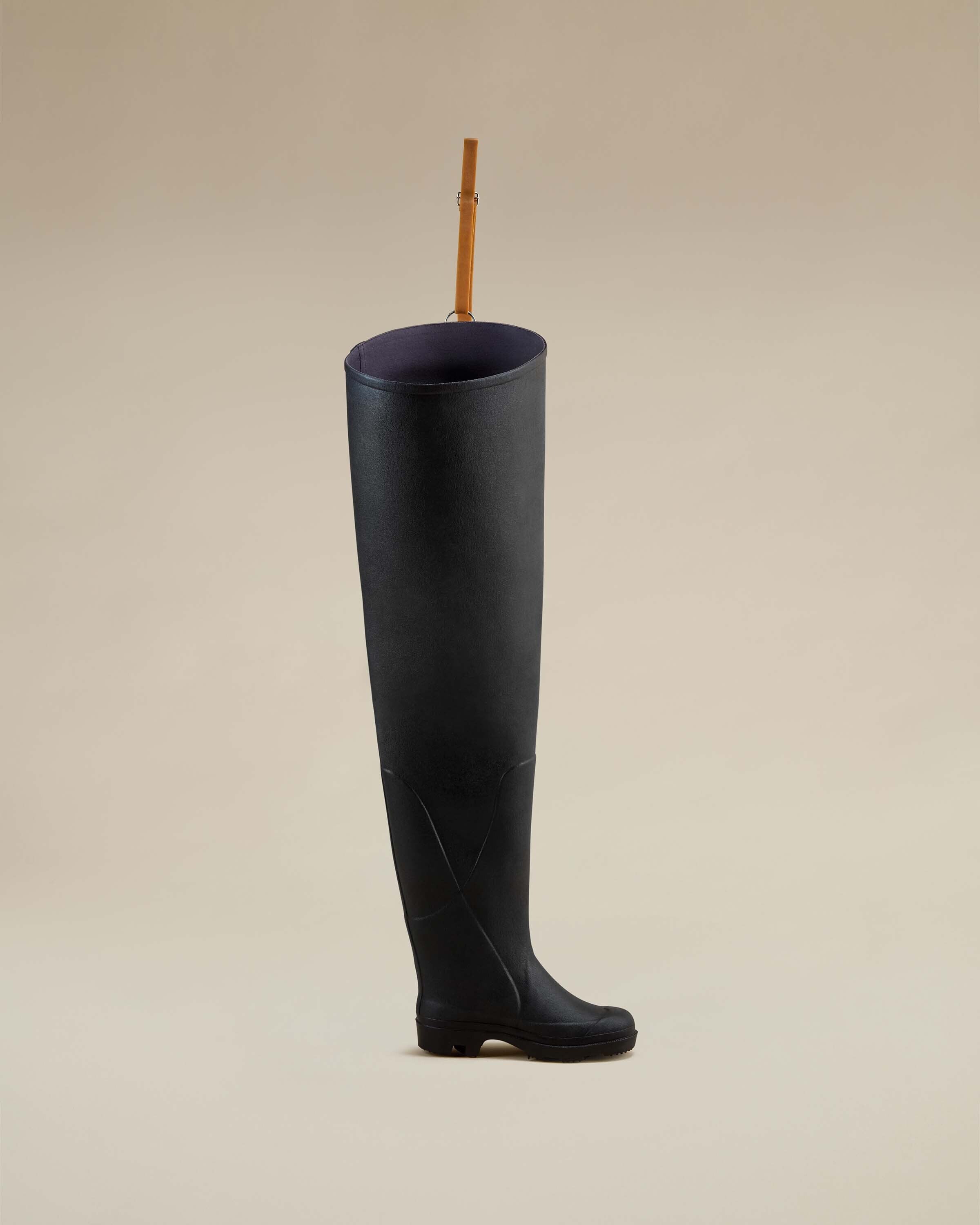 A Women's Océane Ponti Lined Thigh Boot in Black displayed in a soft beige studio setting, revealing the boot's subtle texture and structured upper, with an upward-facing view of the interior and brown leather suspension strap.