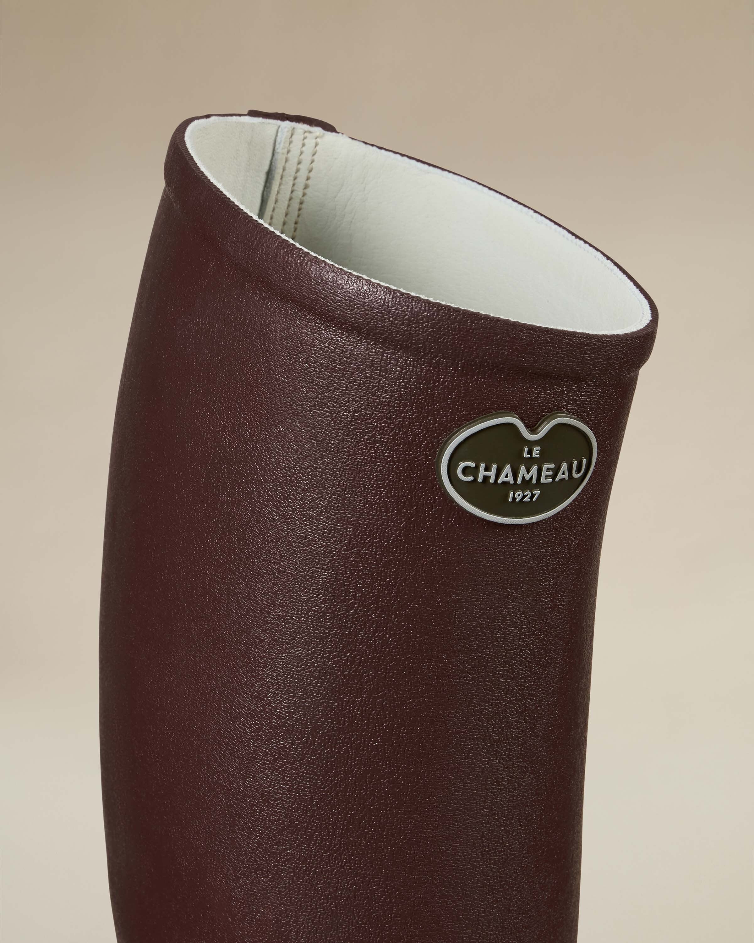 A detailed close-up of the Women's Saint Hubert Leather Lined Wellington Boot in Brown’s upper shaft showing the maroon brown exterior and white leather lining. Prominently displayed is the oval-shaped Le Chameau badge in silver with black lettering that reads “Le Chameau 1927.”
