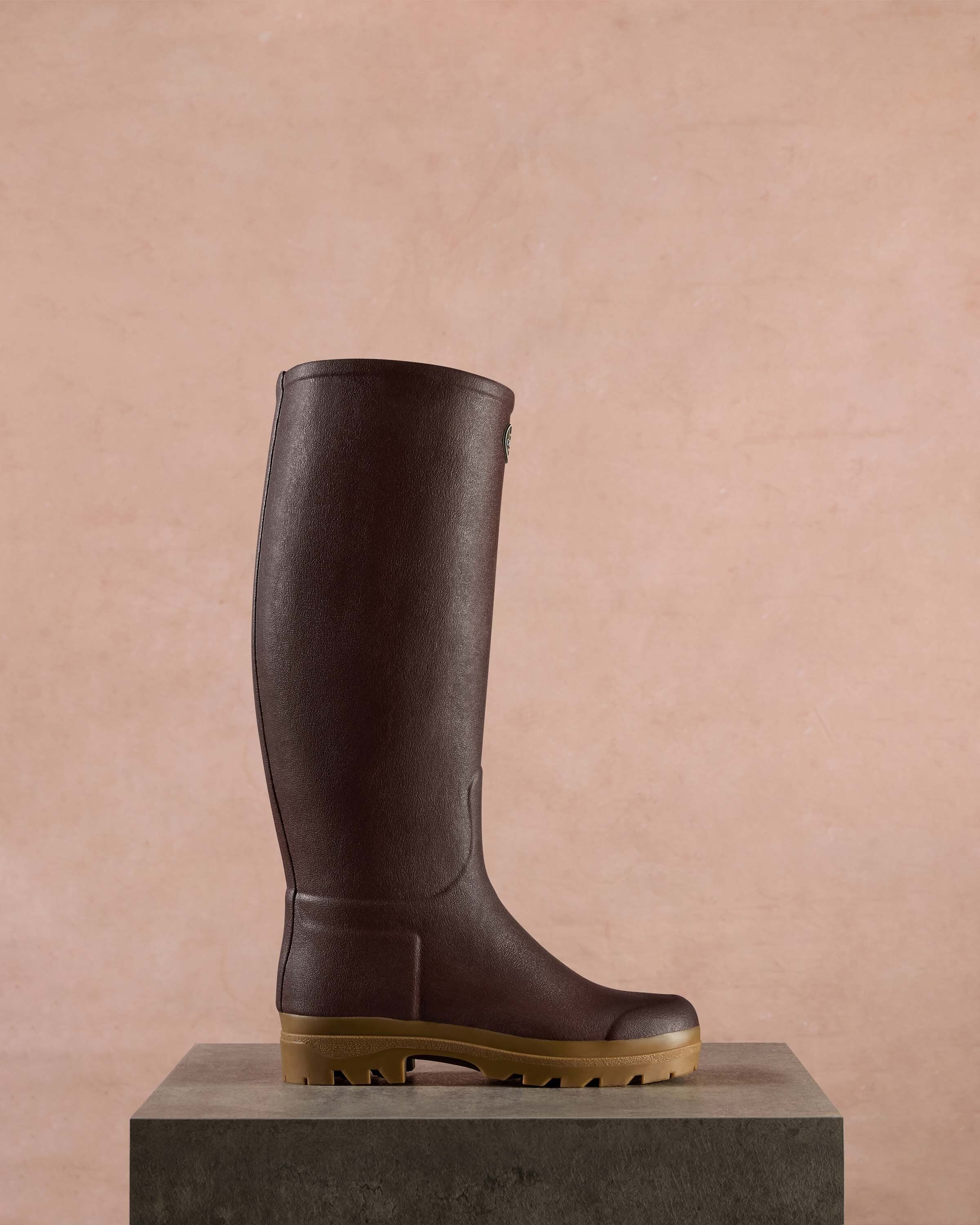 A single Women's Saint Hubert Leather Lined Wellington Boot in Brown shown in profile on a stone-textured pedestal, set against a soft pink background. The clean presentation highlights the boot’s height, smooth finish, and robust gum sole designed for durability and grip.