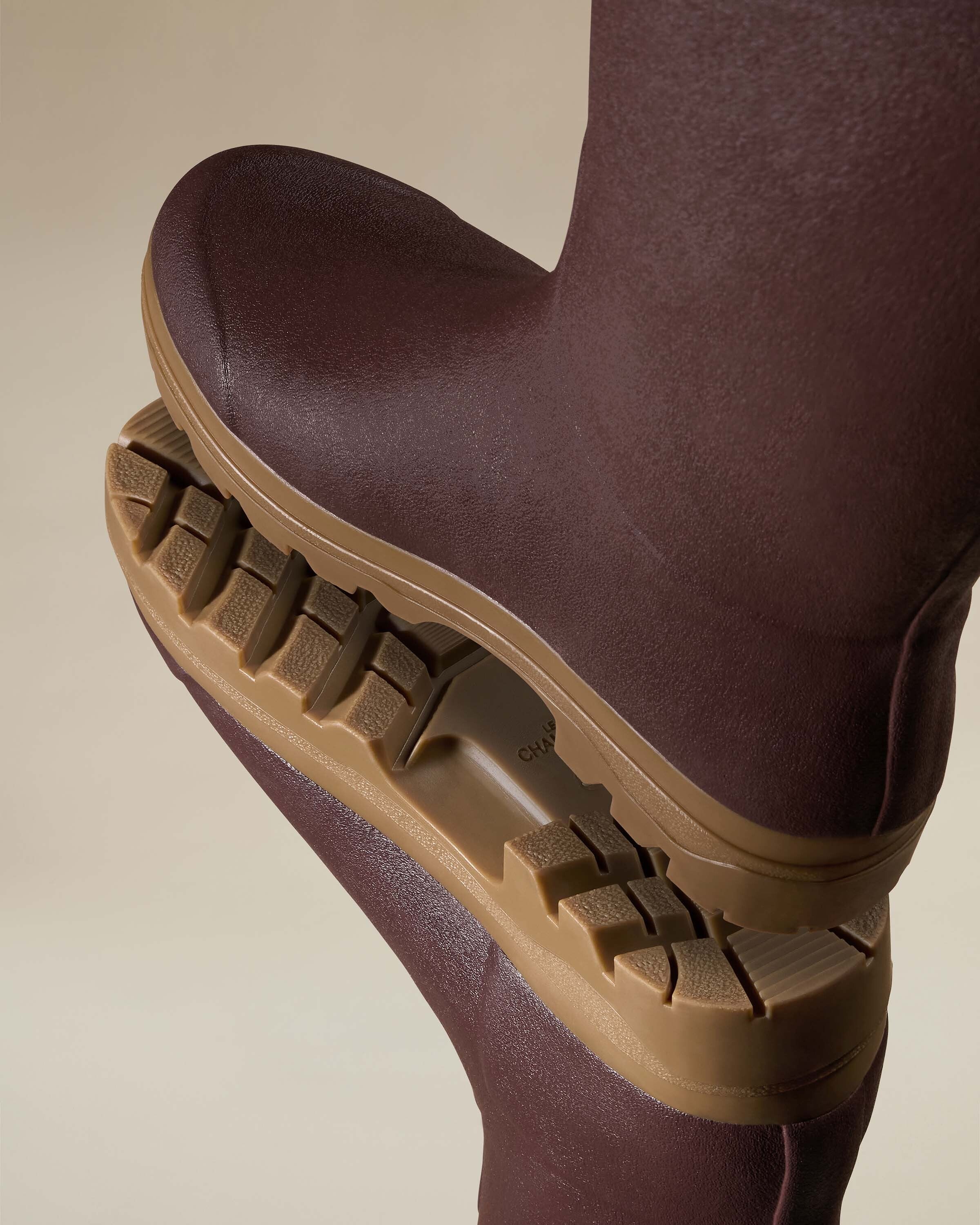 A dynamic close-up of two stacked Women's Saint Hubert Leather Lined Wellington Boot in Brown with one showing the sole. The thick gum rubber sole has deep lugs and aggressive treads for traction. The image emphasises the boot's rugged construction and outdoor performance capability.