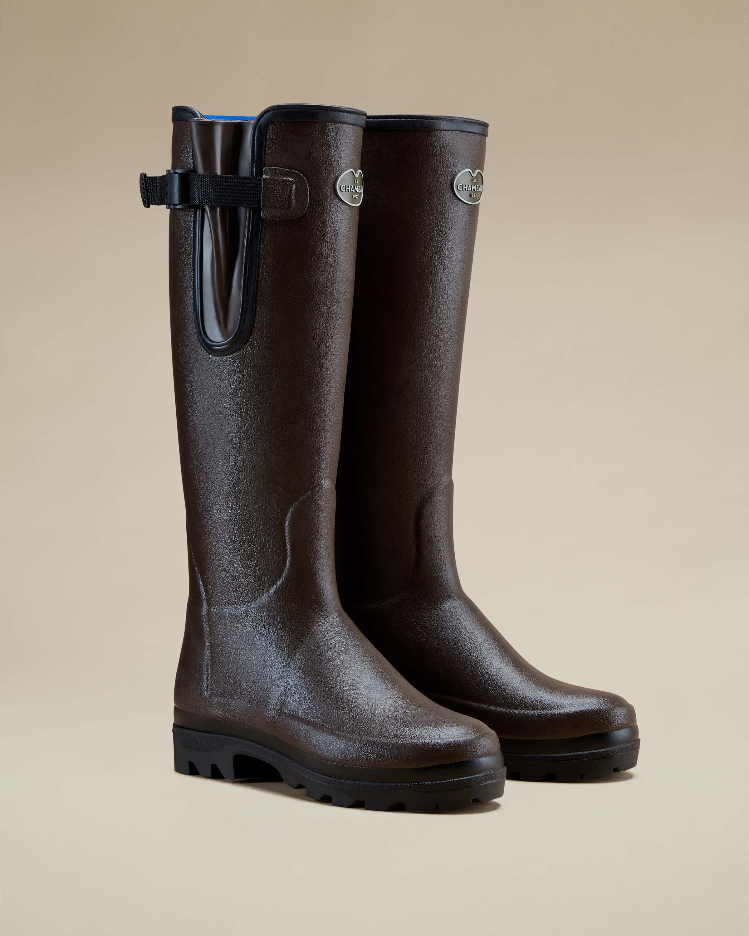 A pair of Women's Vierzonord Neoprene Lined Wellington Boot in Dark Brown positioned at a 45-degree angle on a neutral tan backdrop. The boots stand upright, with one slightly forward, showcasing the smooth, contoured lines of the design. Notable details include the brand’s silver oval logo badge on each shaft, black piping along the top line, a high-grip outsole, and ergonomic shaping that hugs the natural curves of the leg and foot.