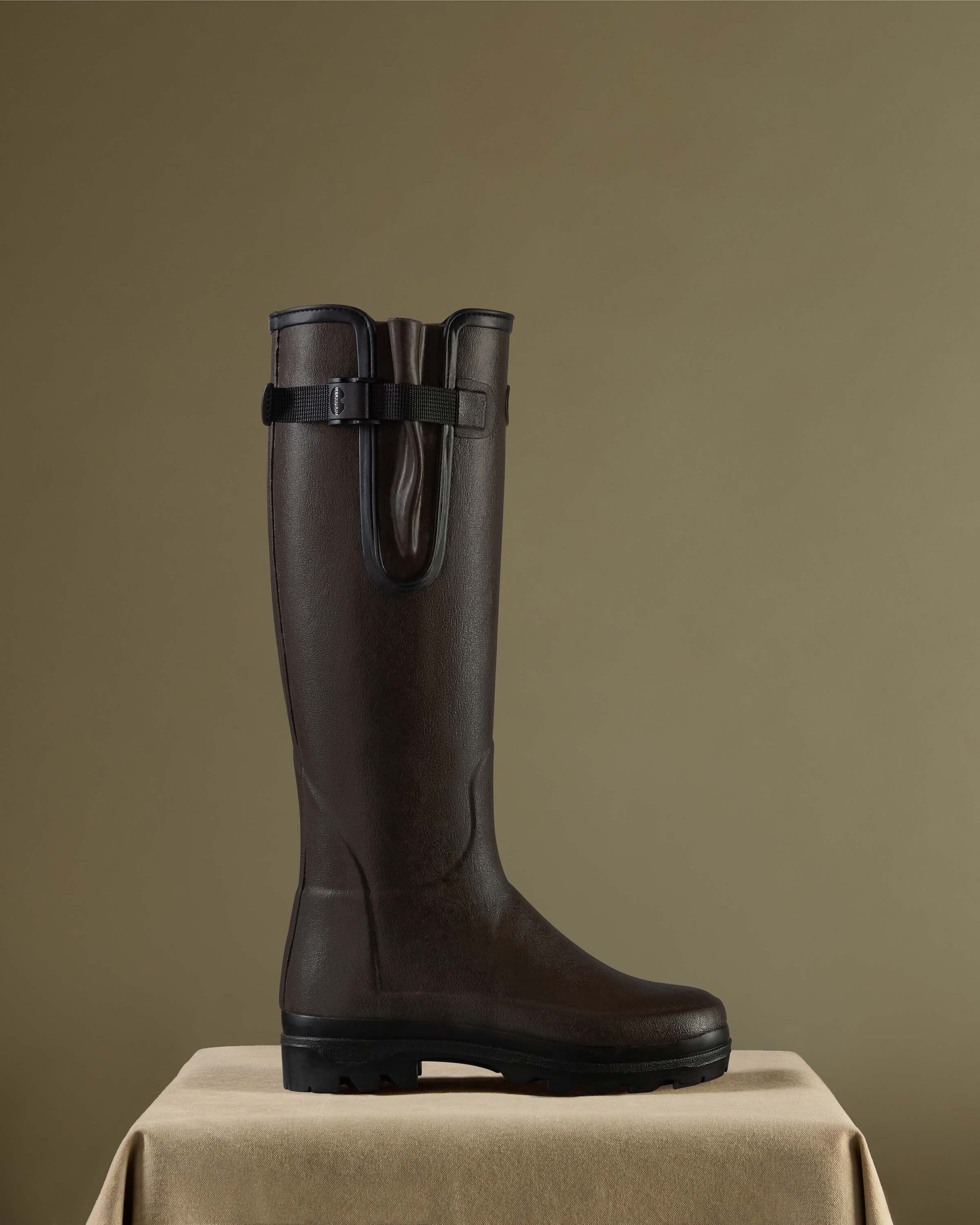 A single Women's Vierzonord Neoprene Lined Wellington Boot in Dark Brown is elegantly displayed on a tan velvet-covered pedestal against a warm olive background. The boot is photographed in profile to highlight the sleek silhouette, the subtle sheen of the rubber surface, and the side gusset design. The presentation evokes premium craftsmanship and traditional French outdoor style.