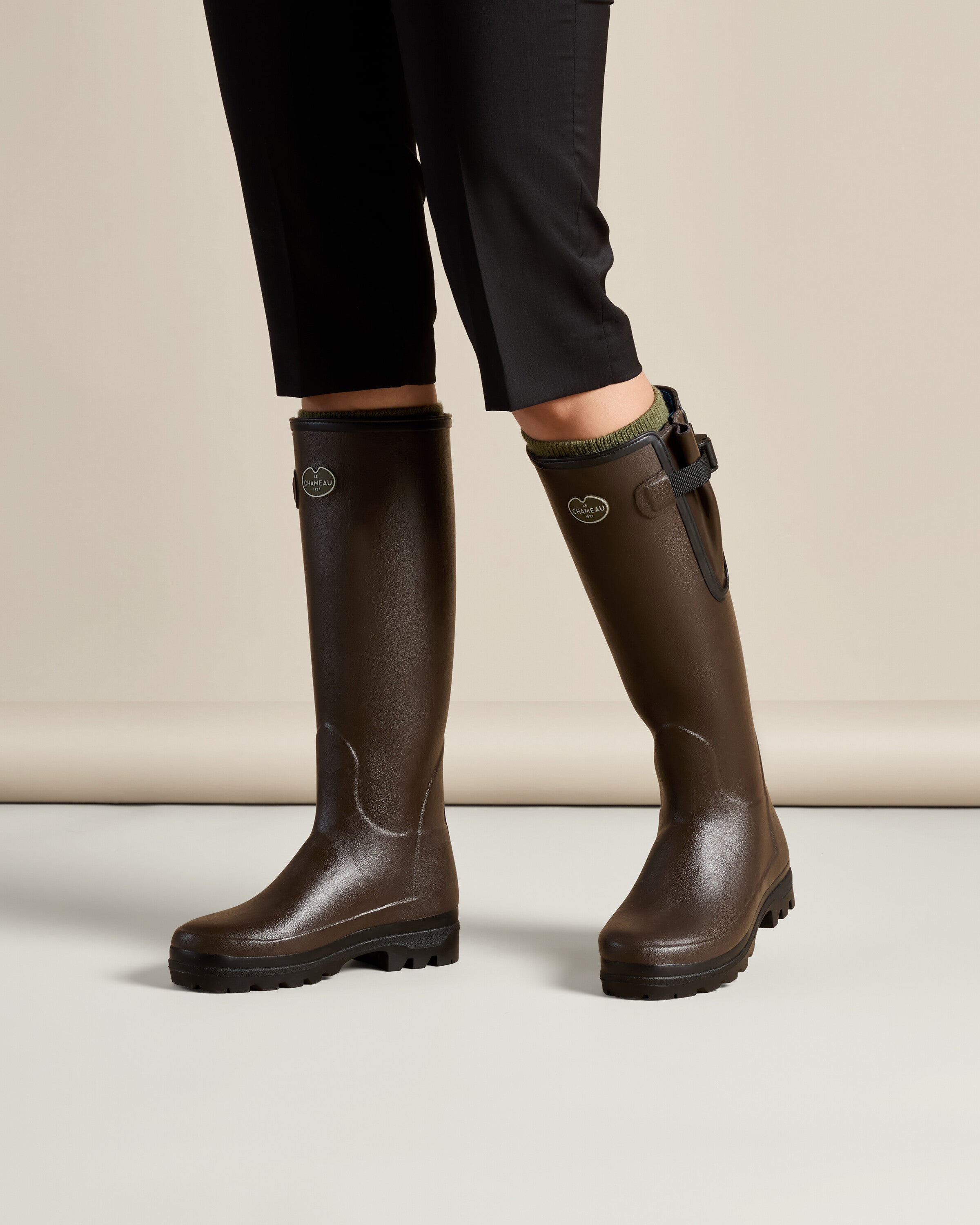 A lifestyle image of a person wearing the Women's Vierzonord Neoprene Lined Wellington Boot in Dark Brown paired with cropped black trousers and green socks. The person is standing with feet apart on a studio floor, offering a realistic look at how the boots fit snugly around the lower leg. The soft neoprene lining is slightly visible, adding a pop of color and warmth.