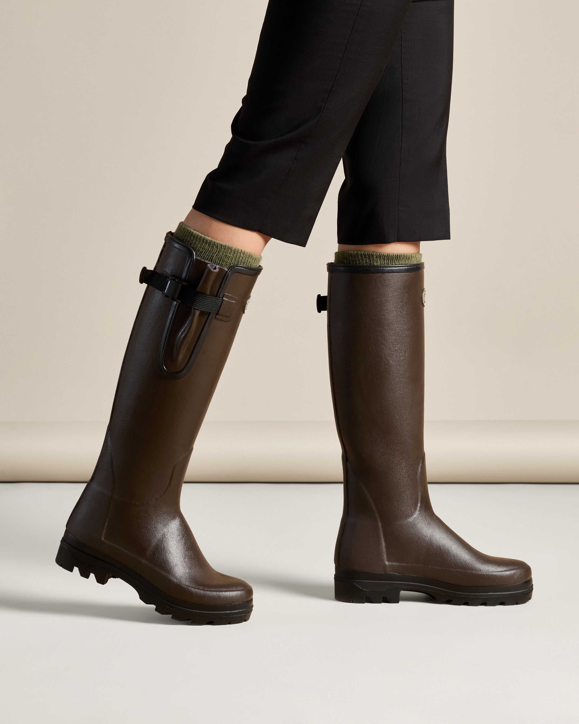Side profile view of a model mid-stride wearing the Women's Vierzonord Neoprene Lined Wellington Boot in Dark Brown, revealing the natural motion and ergonomic shaping of the boot as the front leg bends slightly forward. The adjustable strap and gusset are clearly visible, and the rugged sole flexes with the step, illustrating the boot’s comfort and mobility for extended use outdoors.