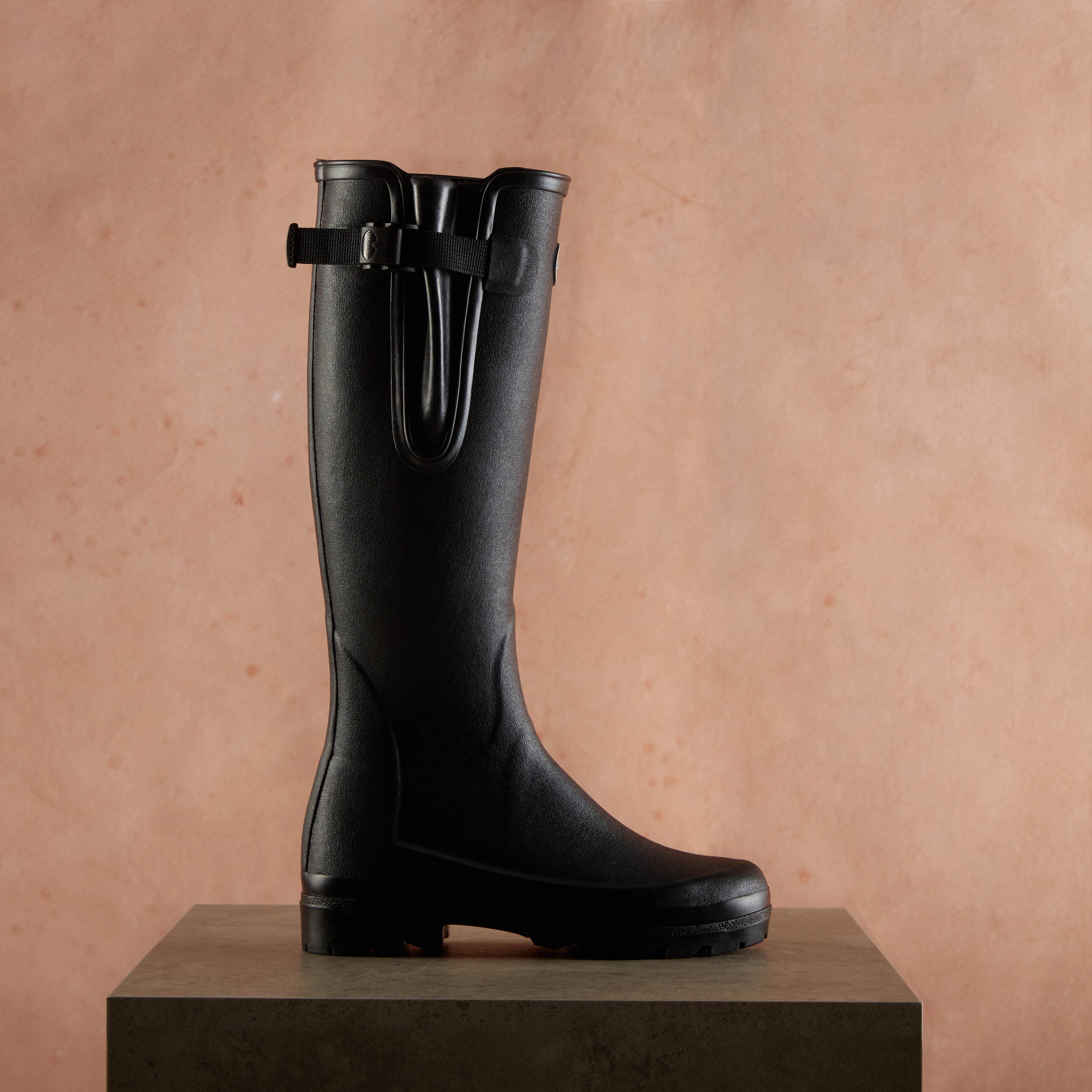 Women's Vierzonord Neoprene Lined Wellington Boot in Black displayed upright on a concrete pedestal against a peach-toned background, highlighting the boot’s ergonomic shape and high leg cut.