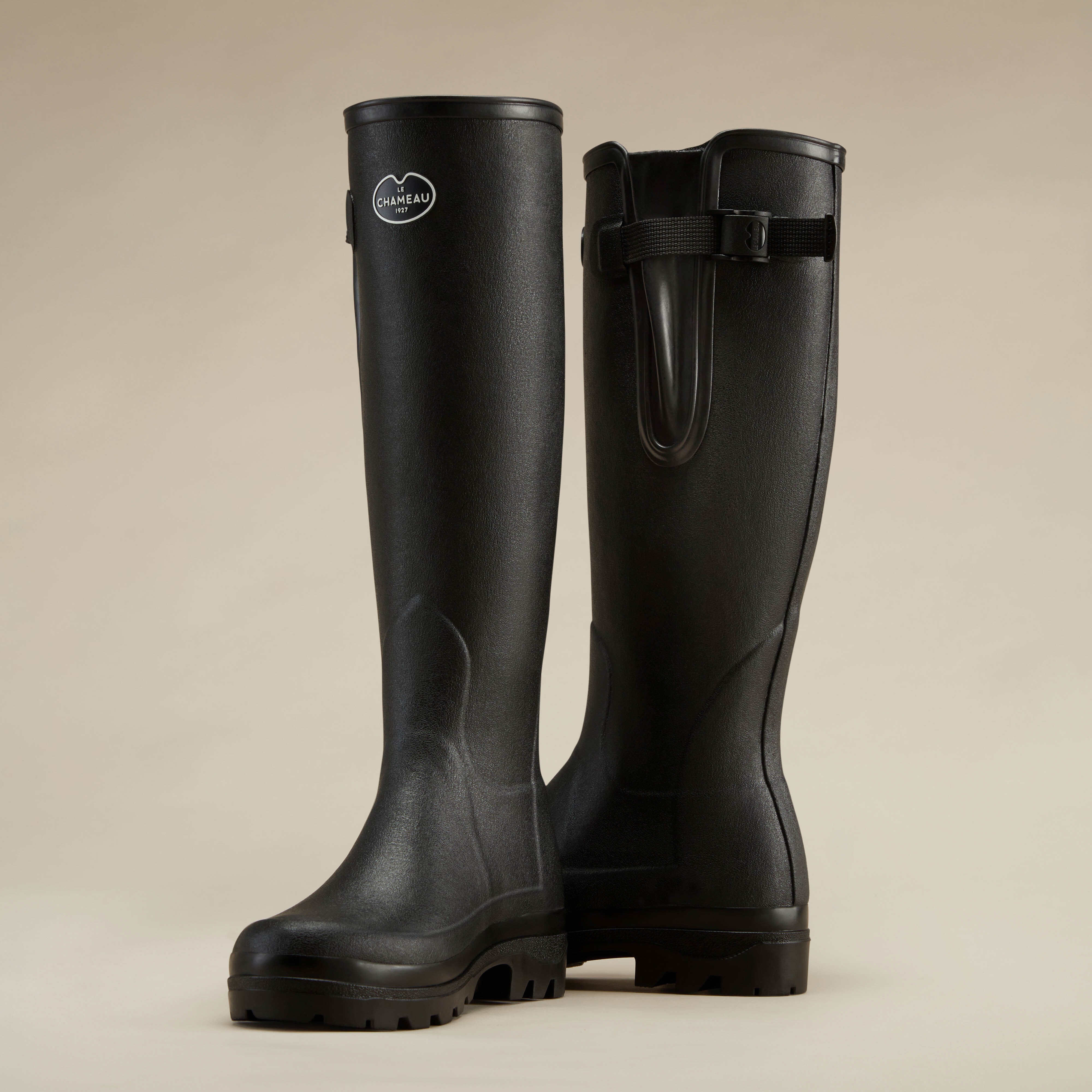 Stylized silhouette shot of two Women's Vierzonord Neoprene Lined Wellington Boot in Black, viewed from the side on a beige surface, focusing on their tall structure and buckle detailing.