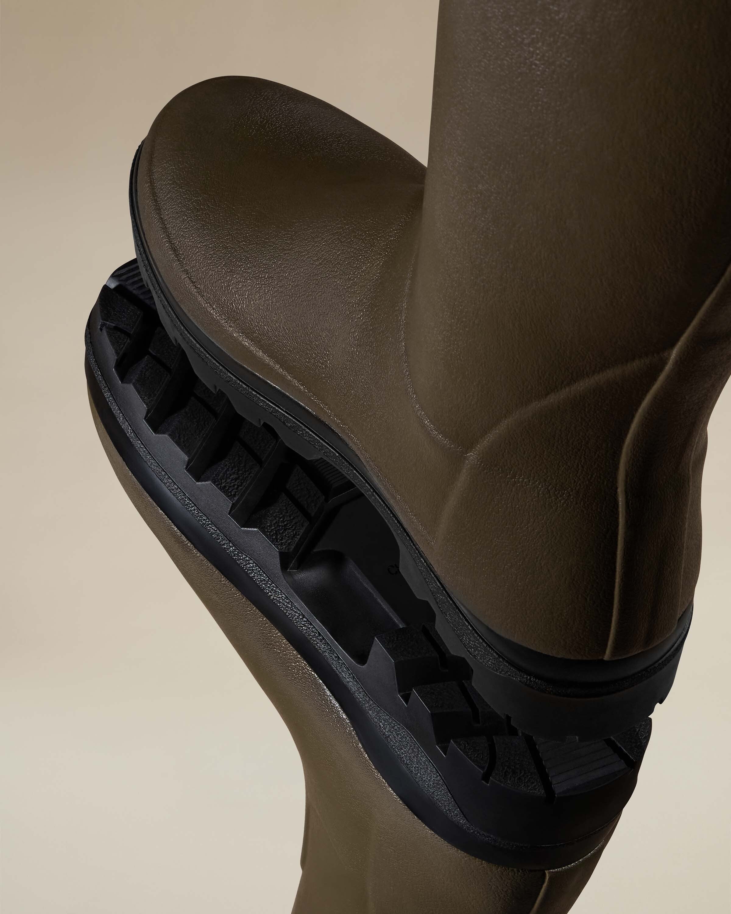 Dramatic overhead shot of one Women's Vierzonord Neoprene Lined Wellington Boot in Chameau Green laid over another, focusing on the heavy-duty, multi-directional lug sole. The rugged rubber outsole and arch-support structure are emphasised in the soft lighting.