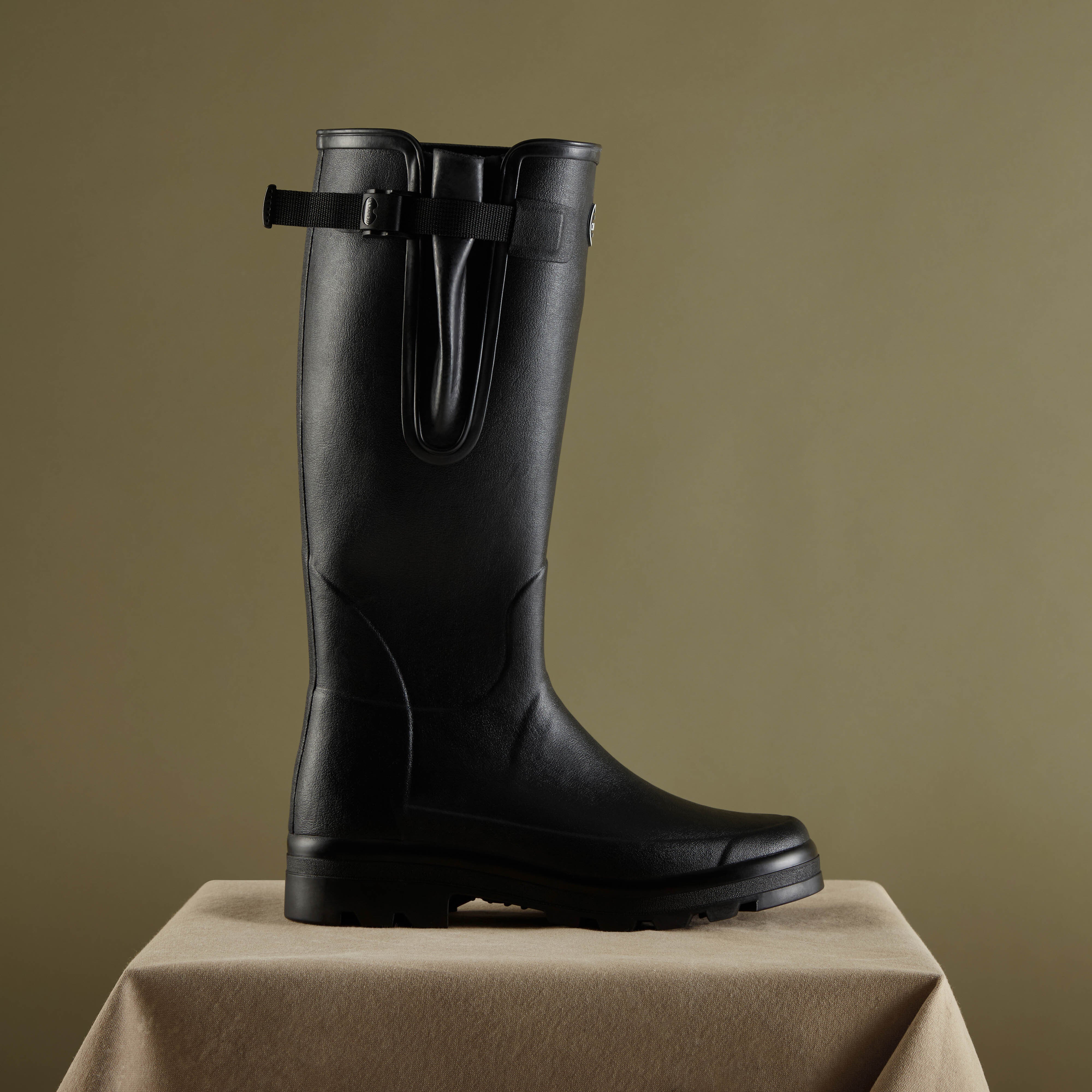 Artistic side profile of the same Men's Vierzonord Neoprene Lined Wellington Boot in Black on a beige pedestal, photographed against a muted olive background. The texture and craftsmanship of the boot's rubber exterior are highlighted by soft directional lighting.