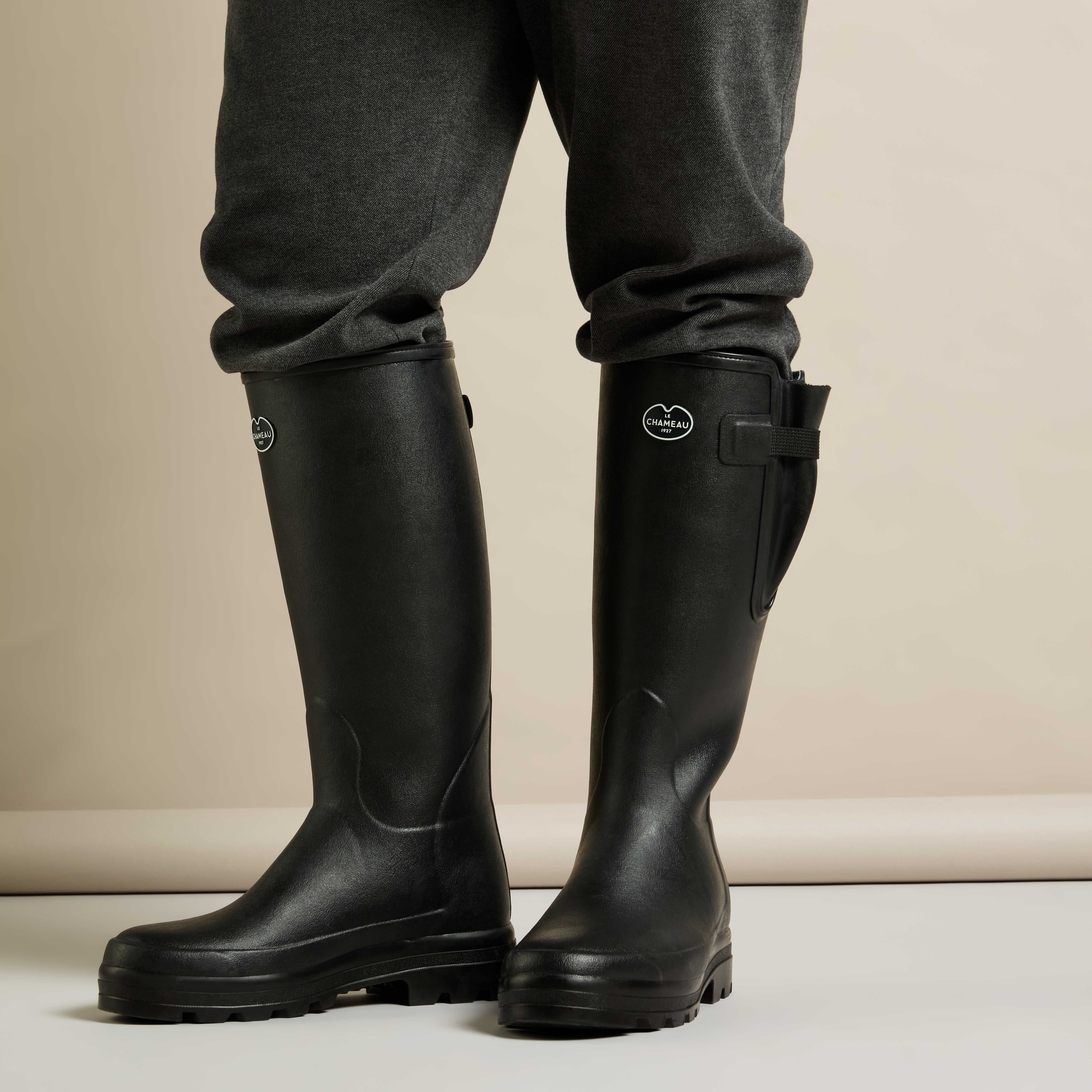 Model standing in the Men's Vierzonord Neoprene Lined Wellington Boot in Black against a light beige backdrop, wearing charcoal grey trousers tucked into the boots. This image highlights the fit and form of the boots when worn, as well as the contrast between the boot and the outfit.