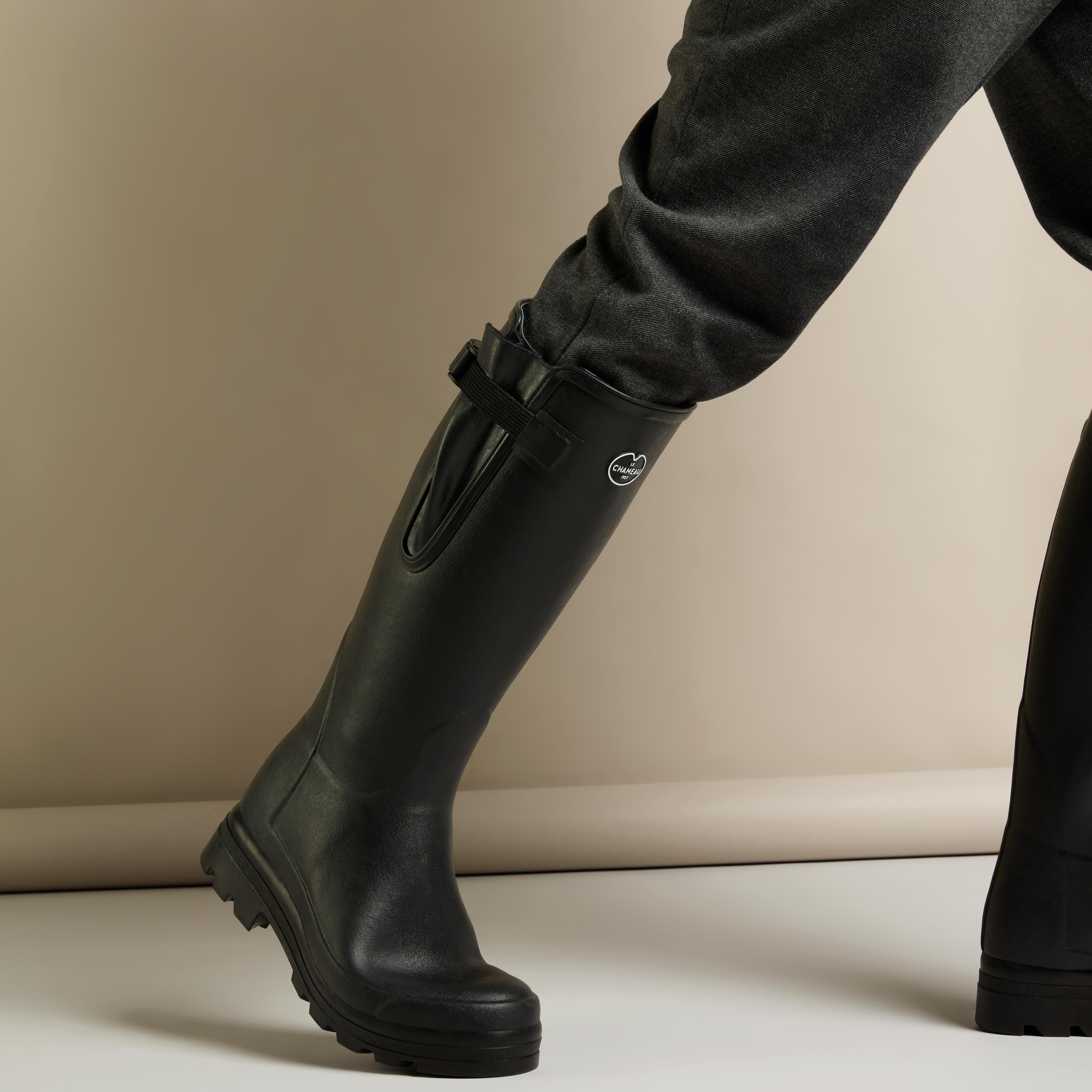 Side walking shot of the model lifting one foot mid-step, showing boot flexibility and ankle movement. The adjustable gusset is visible, and the trousers remain tucked neatly into the Men's Vierzonord Neoprene Lined Wellington Boot in Black, reinforcing their functional design.