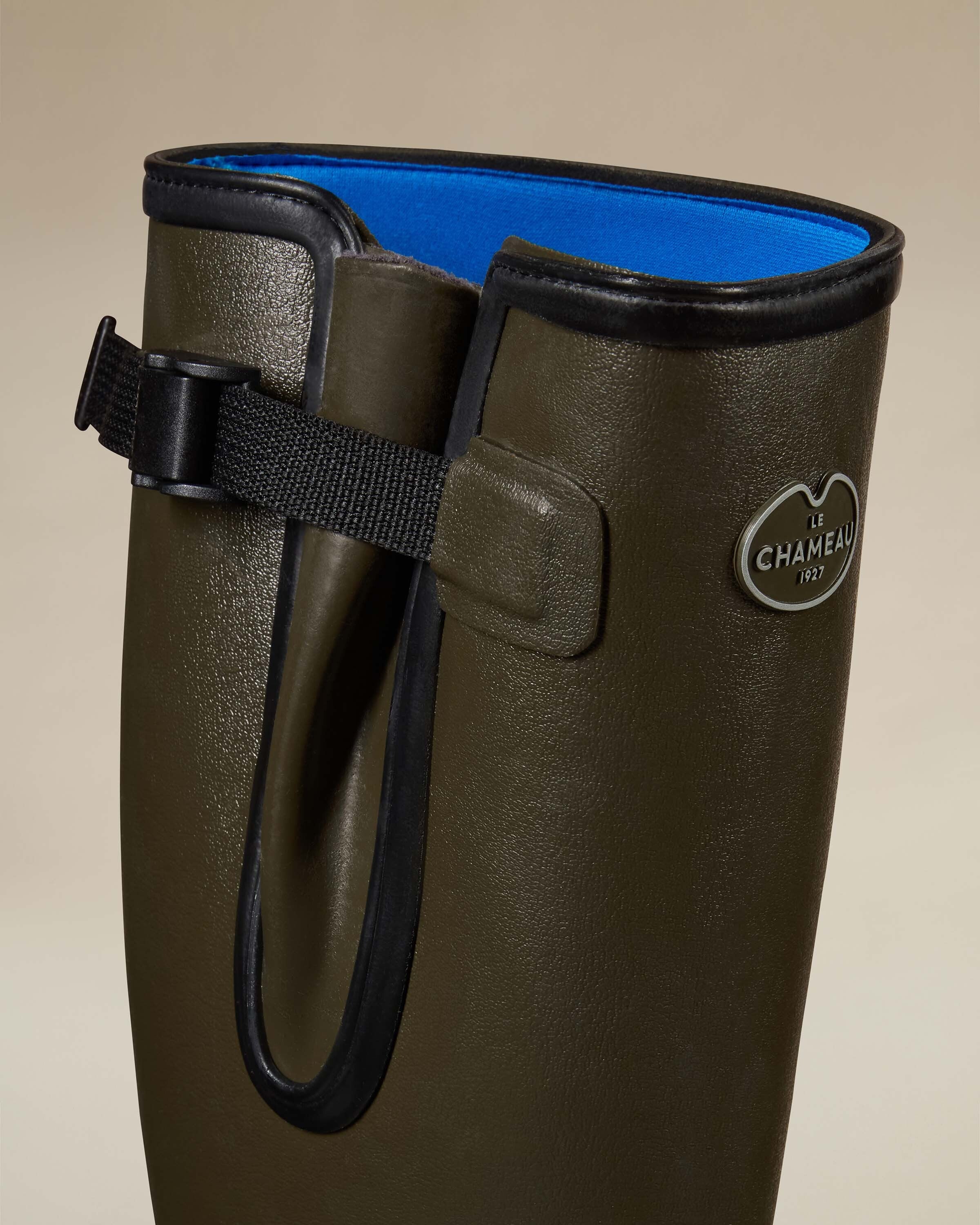 Close-up detail of the upper shaft on the Men's Vierzonord Neoprene Lined Wellington Boot in Chameau Green. The photo showcases the textured green rubber surface, black-edged gusset, and the brand’s silver badge logo with "Le Chameau 1927" engraved. The bright blue neoprene lining is partially visible inside the collar.