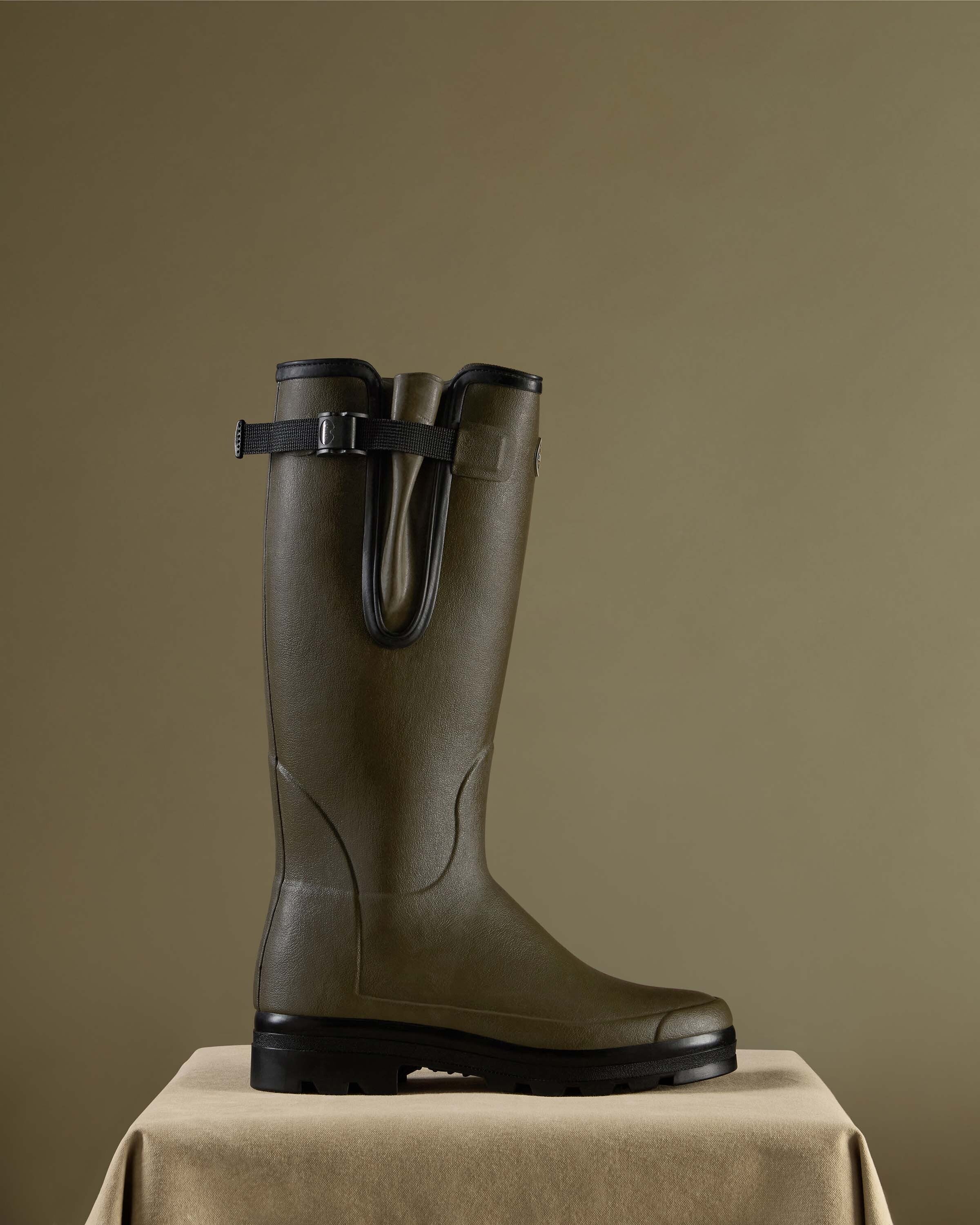 A single Men's Vierzonord Neoprene Lined Wellington Boot in Chameau Green presented on a tan suede-textured pedestal against an olive-colored backdrop. The image emphasises the tall, structured shaft and signature side gusset with black trim, illustrating the boot’s premium construction and outdoor utility.