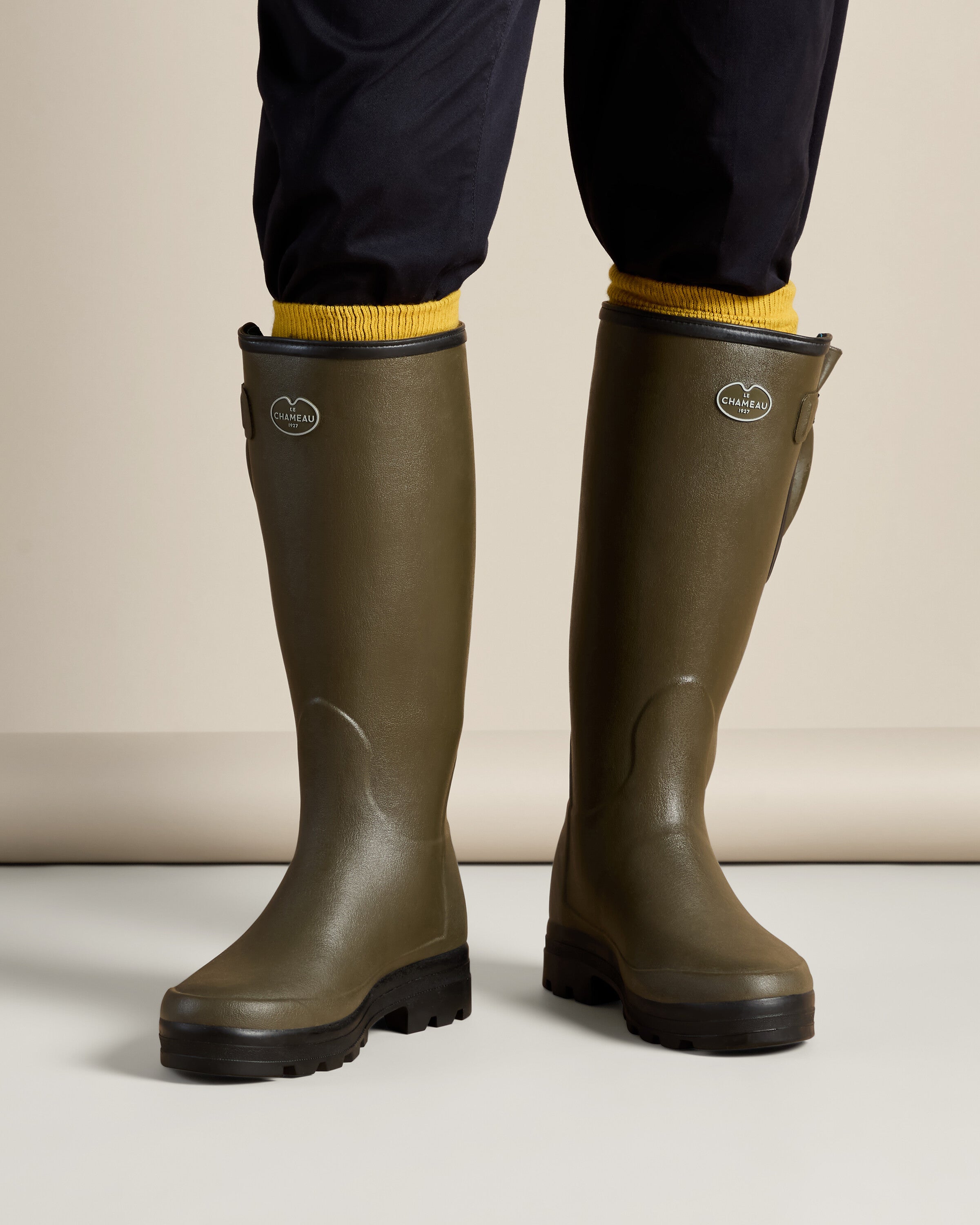 On-foot front view of a model wearing the Men's Vierzonord Neoprene Lined Wellington Boot in Chameau Green, paired with navy blue pants and yellow socks. The image highlights the comfortable fit and stylish contrast between functional outdoor footwear and casual wear.