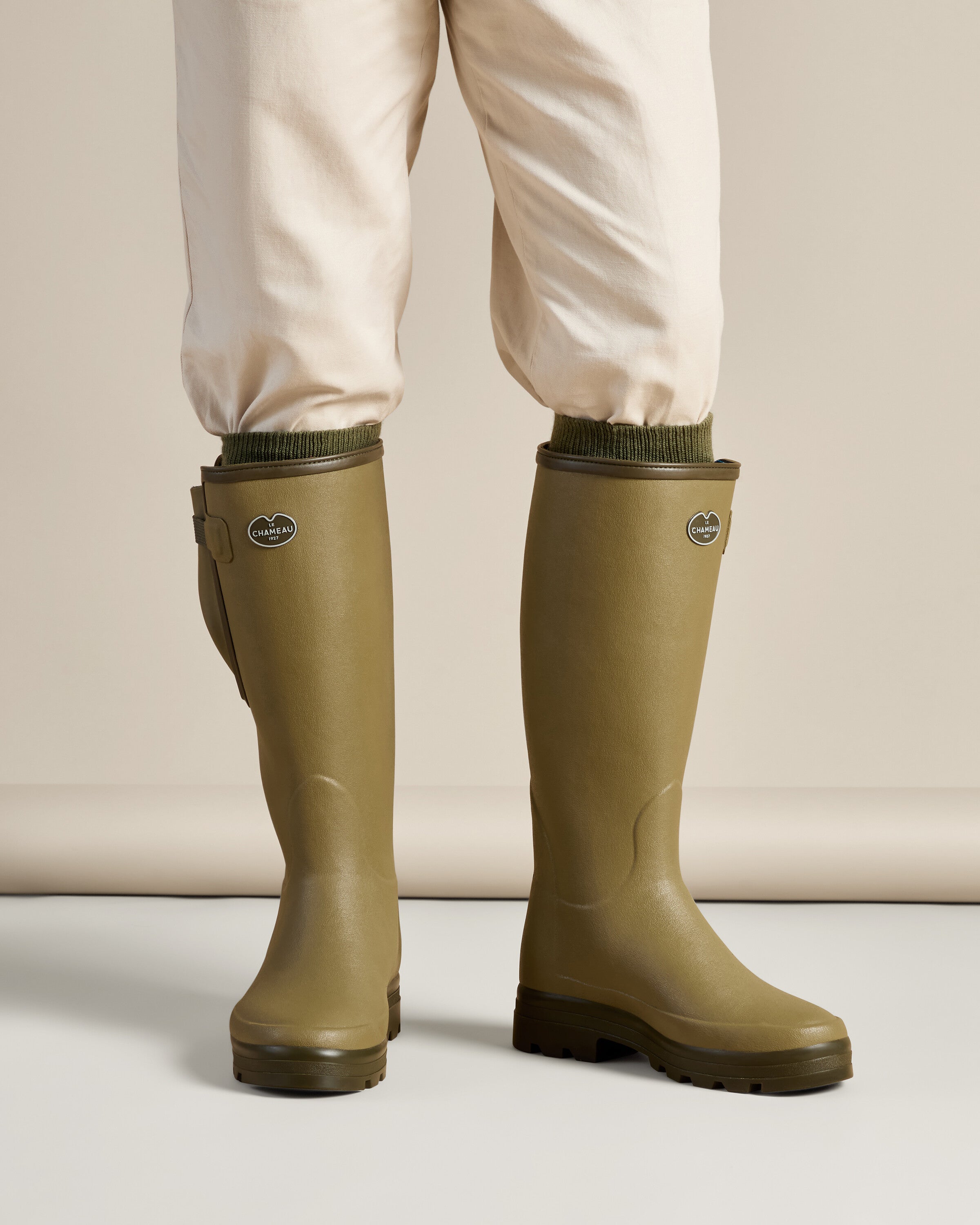 Model wearing the Men's Vierzonord Neoprene Lined Wellington Boot in Iconic Green, standing on a neutral set. The boots rise to just below the knee, worn over light beige trousers tucked in at the cuff, showing a clean and practical outdoor look.