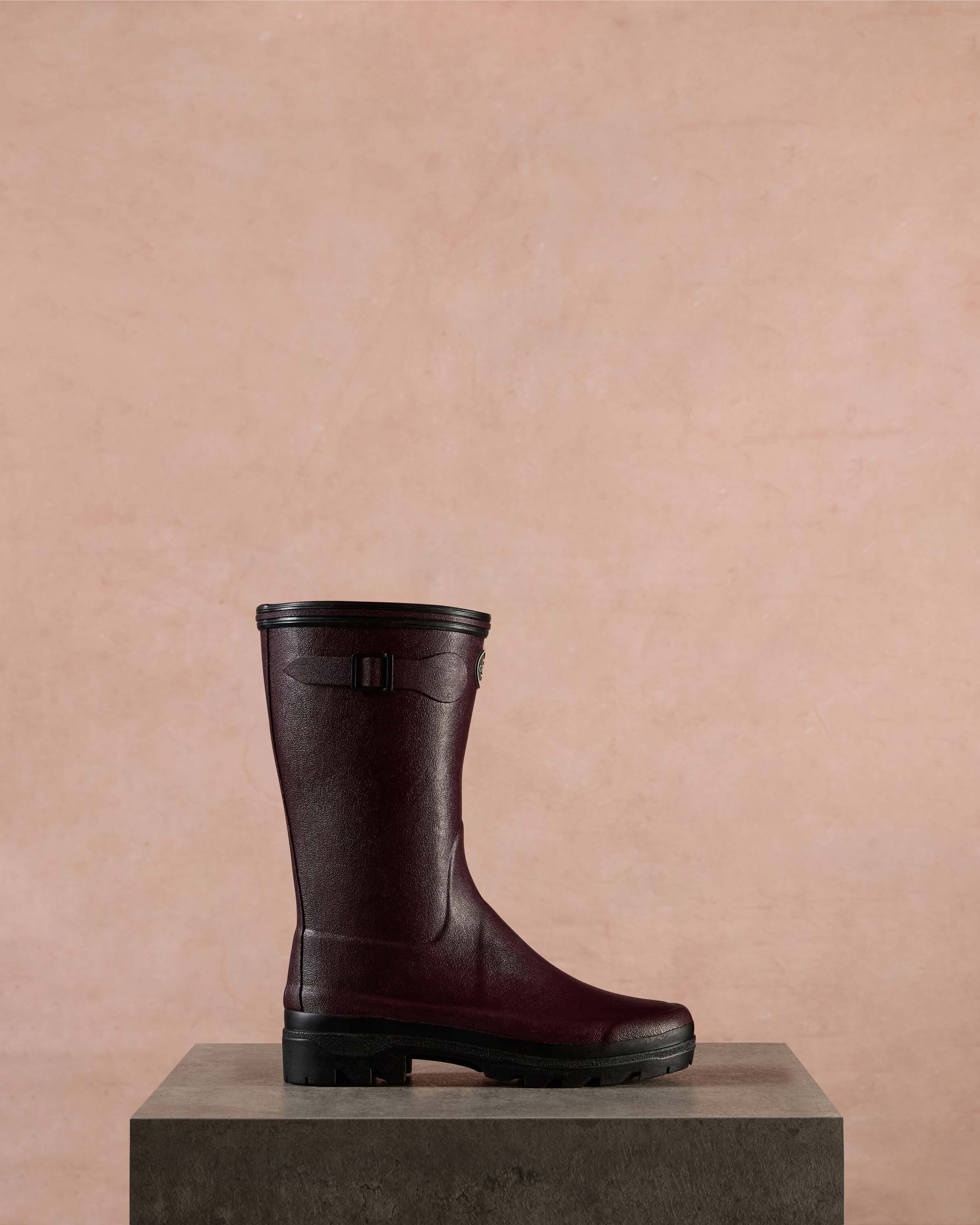 Single Women's Giverny Jersey Lined Bottillon in Cherry Red displayed in profile on a dark stone pedestal against a soft pink background. The boot’s sleek rubber surface, curved seam detail, and thick black sole are clearly emphasised.