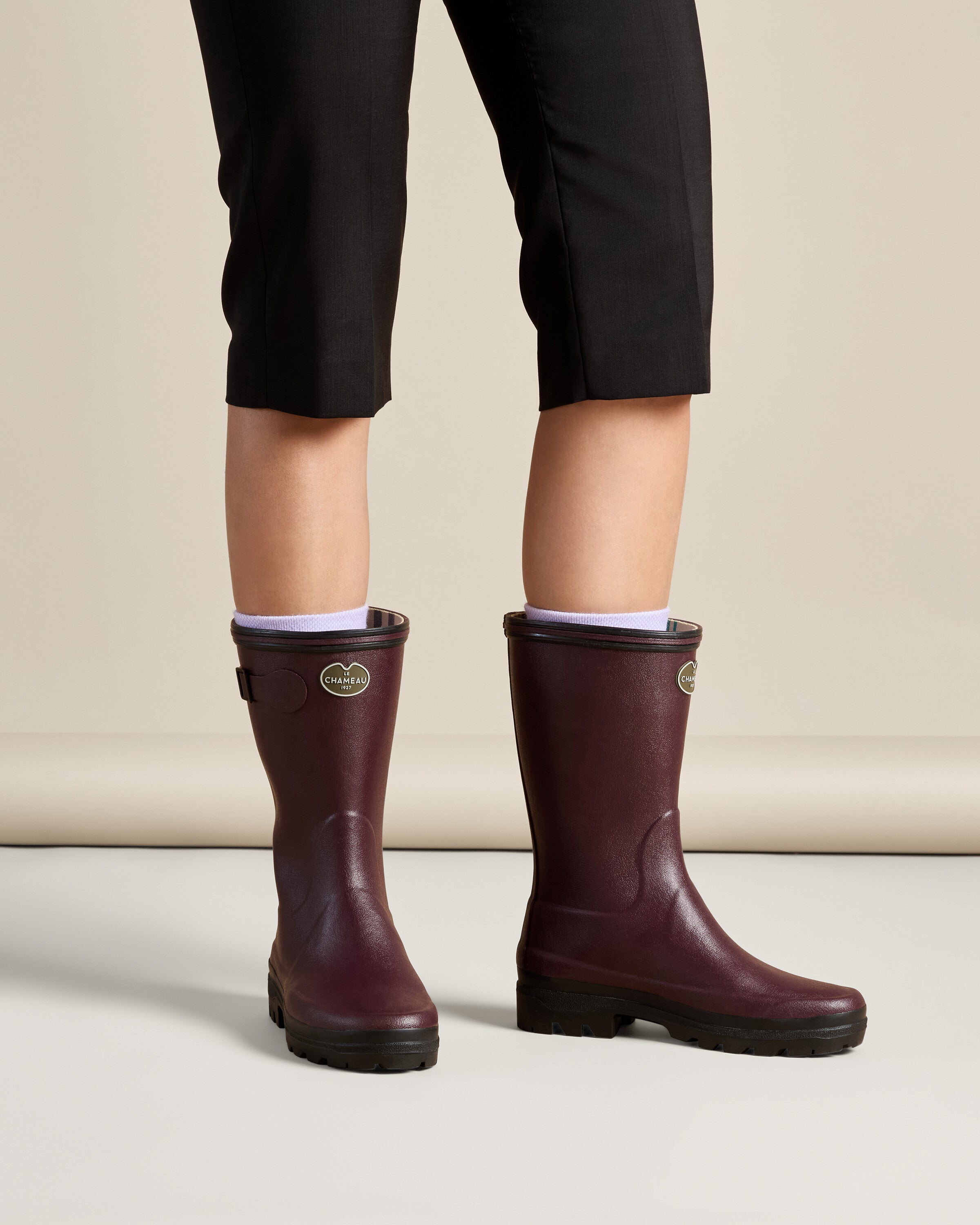 Front-facing view of a person wearing a pair of Women's Giverny Jersey Lined Bottillon in Cherry Red. The model is shown from mid-thigh down, standing on a light background with feet slightly apart. The boots reach mid-calf and have a matte finish, black treaded soles, and subtle branding with a metallic "Le Chameau" badge on the outer shaft. The boots are styled with white socks and black cropped pants, showcasing a practical yet polished look.