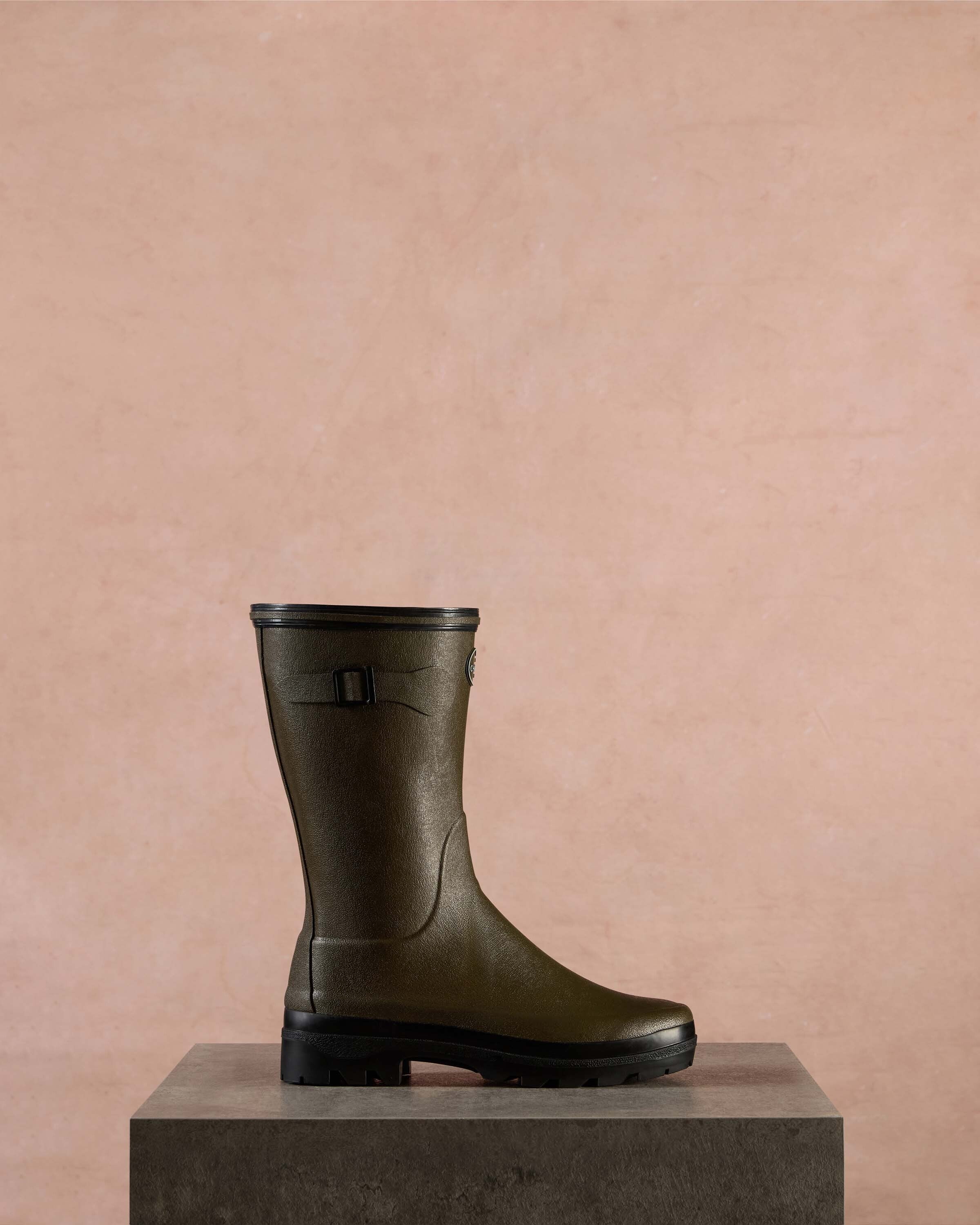 Single Women's Giverny Jersey Lined Bottillon in Chameau Green displayed in profile on a stone pedestal against a muted pink background. The lighting highlights the boot's textured rubber exterior and black tread sole.