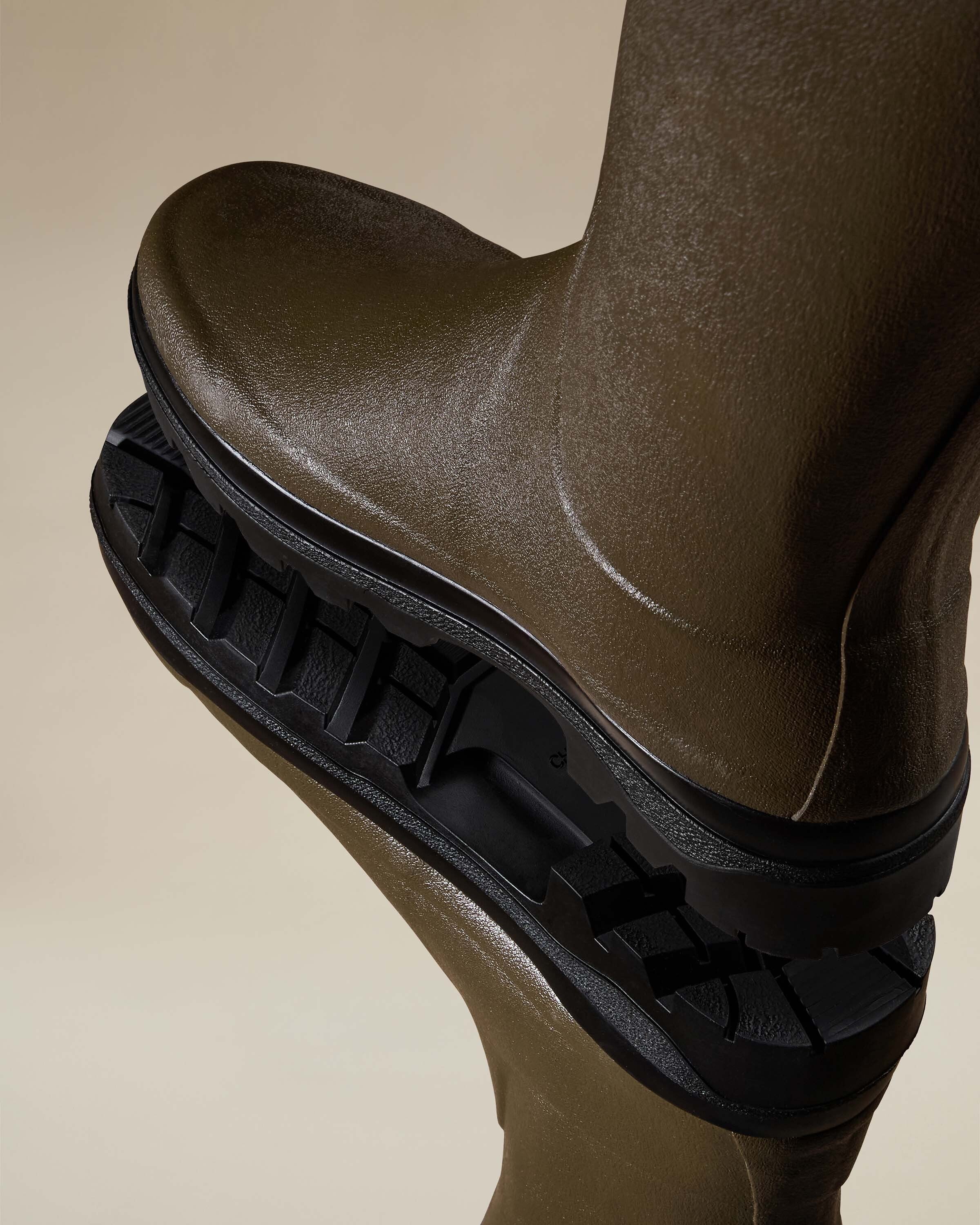 Dynamic close-up showing the sole of one boot resting on the upper part of the other. Highlights include the aggressive tread pattern and the Women's Giverny Jersey Lined Bottillon in Chameau Green’s durable construction, with a strong focus on the texture and craftsmanship.