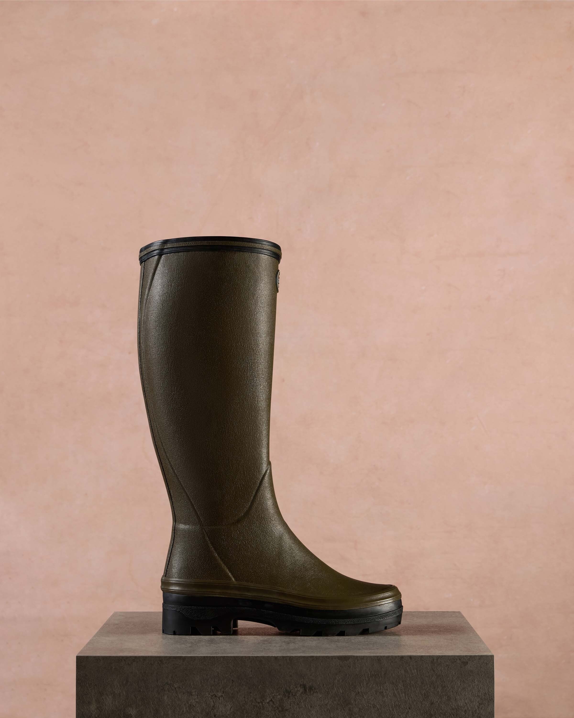 Women's Giverny Jersey Lined Wellington Boot in Chameau Green displayed in profile atop a textured stone pedestal, set against a muted pink-beige background, highlighting the boot’s refined contours and durable construction.