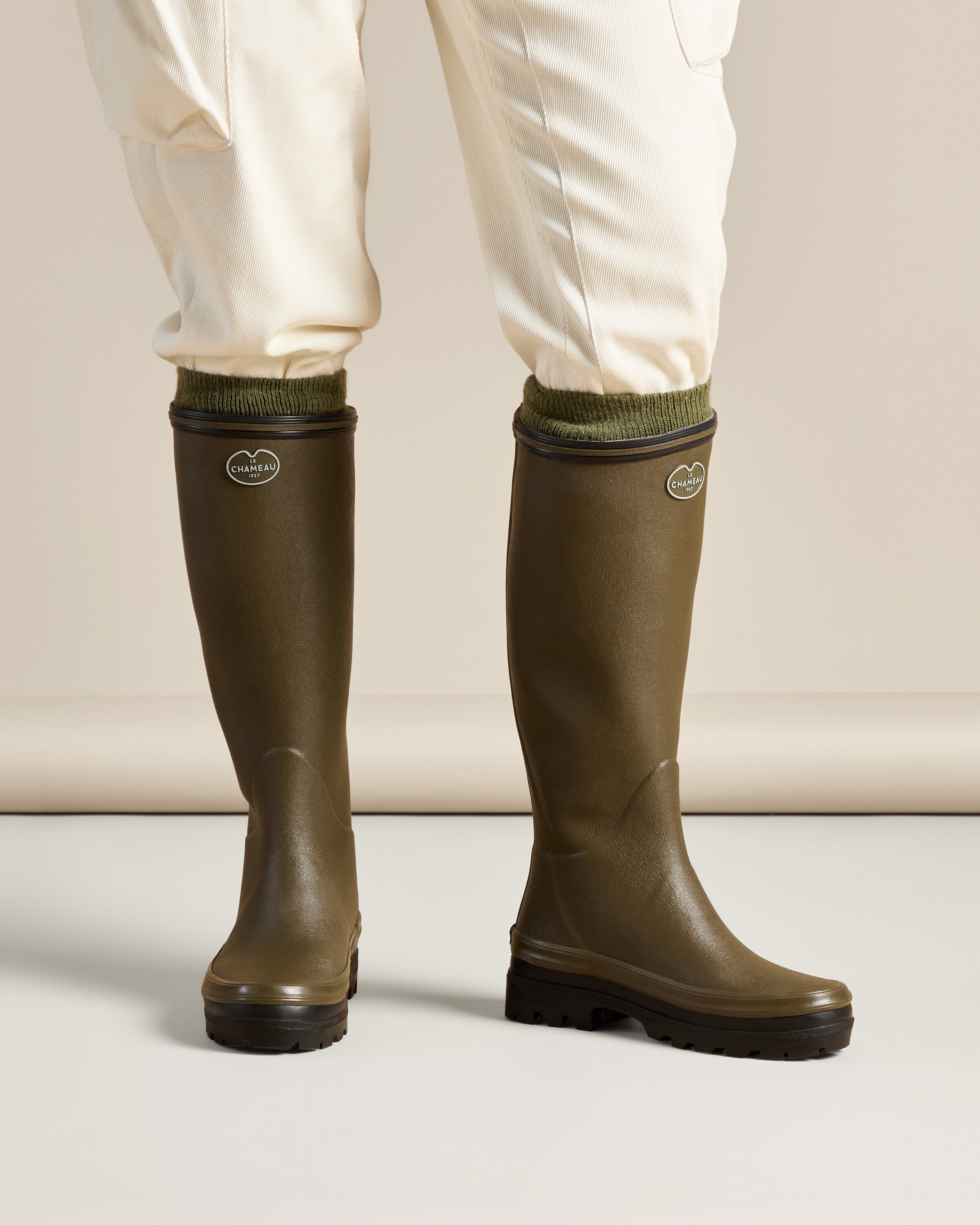 Front-facing view of a model standing in Women's Giverny Jersey Lined Wellington Boot in Chameau Green with light pants tucked in, showcasing the full height and practical fit of the boots in a neutral studio setting.