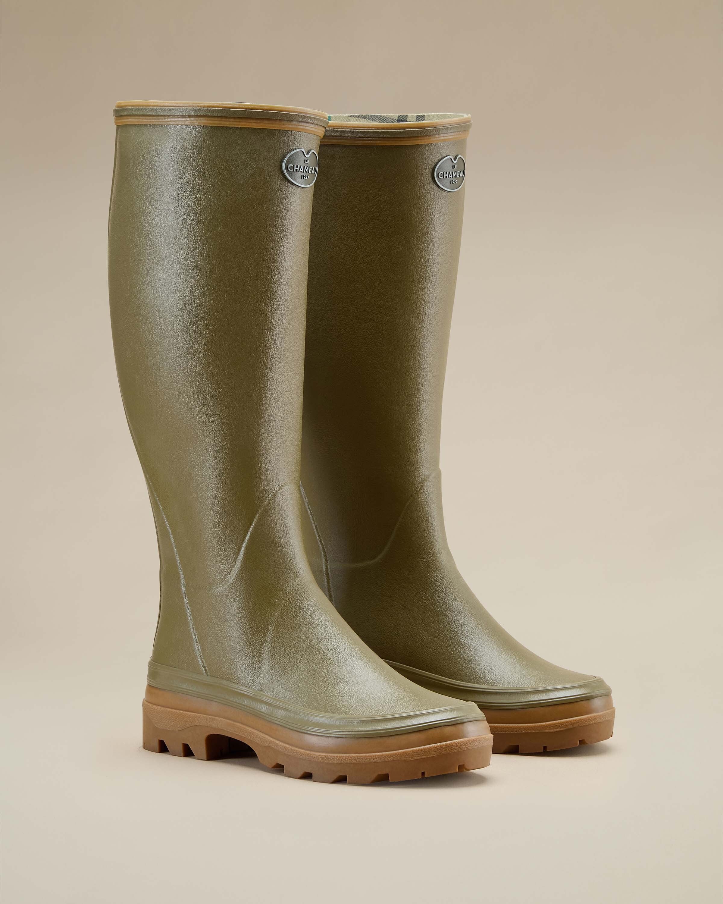 A pair of Women's Giverny Jersey Lined Wellington Boot in Iconic Green is displayed in a three-quarter front view on a beige background. The boots are positioned standing upright, showcasing their contoured shape and structured rubber design. The brown outsole contrasts with the olive green body, and the interior plaid jersey lining is subtly visible.