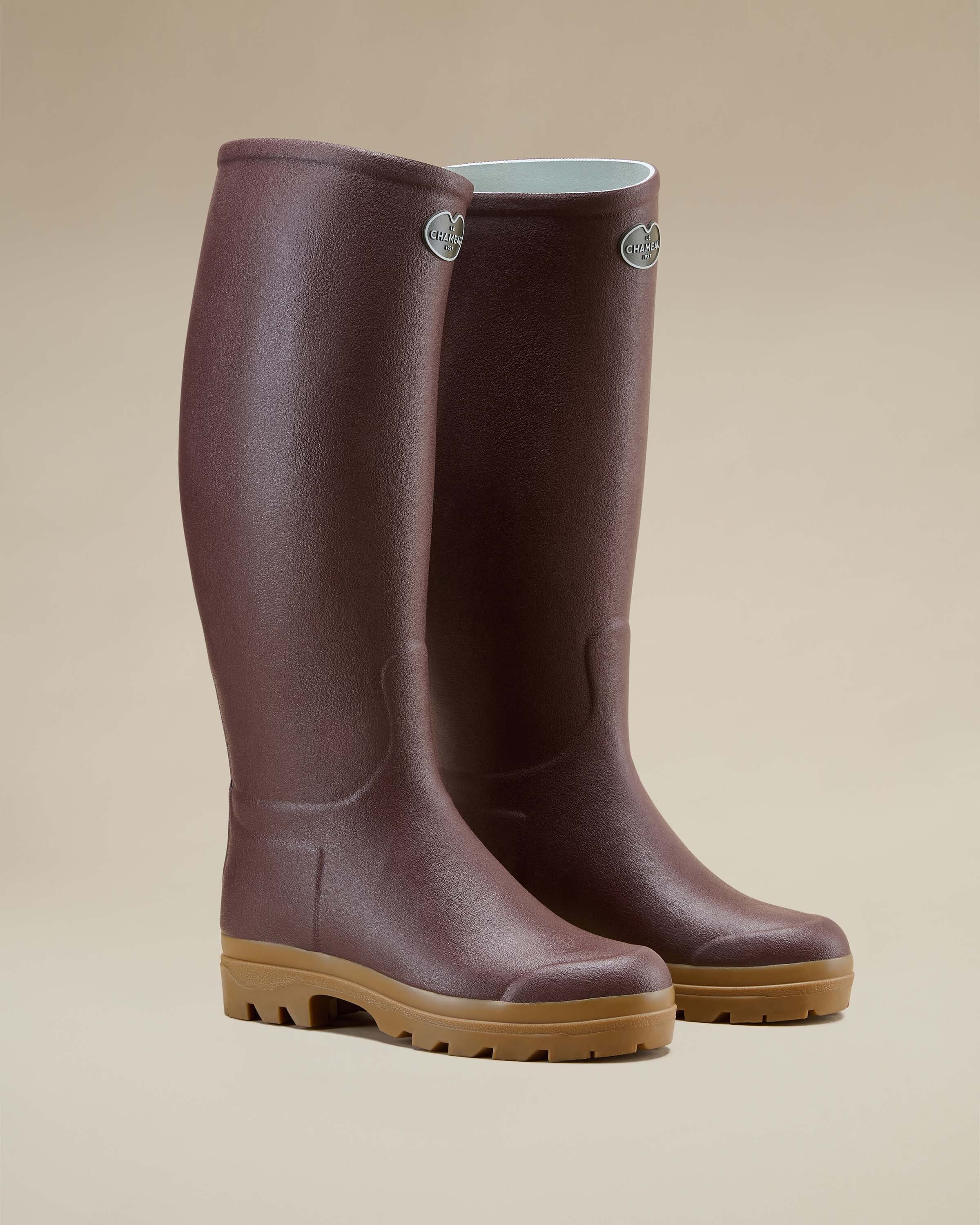 A pair of Women's Saint Hubert Leather Lined Wellington Boot in Brown presented in a three-quarter front view on a neutral beige background. The boots are upright, with one slightly in front of the other, showcasing the brown rubber upper and tan outsole, with Le Chameau logos visible near the top.