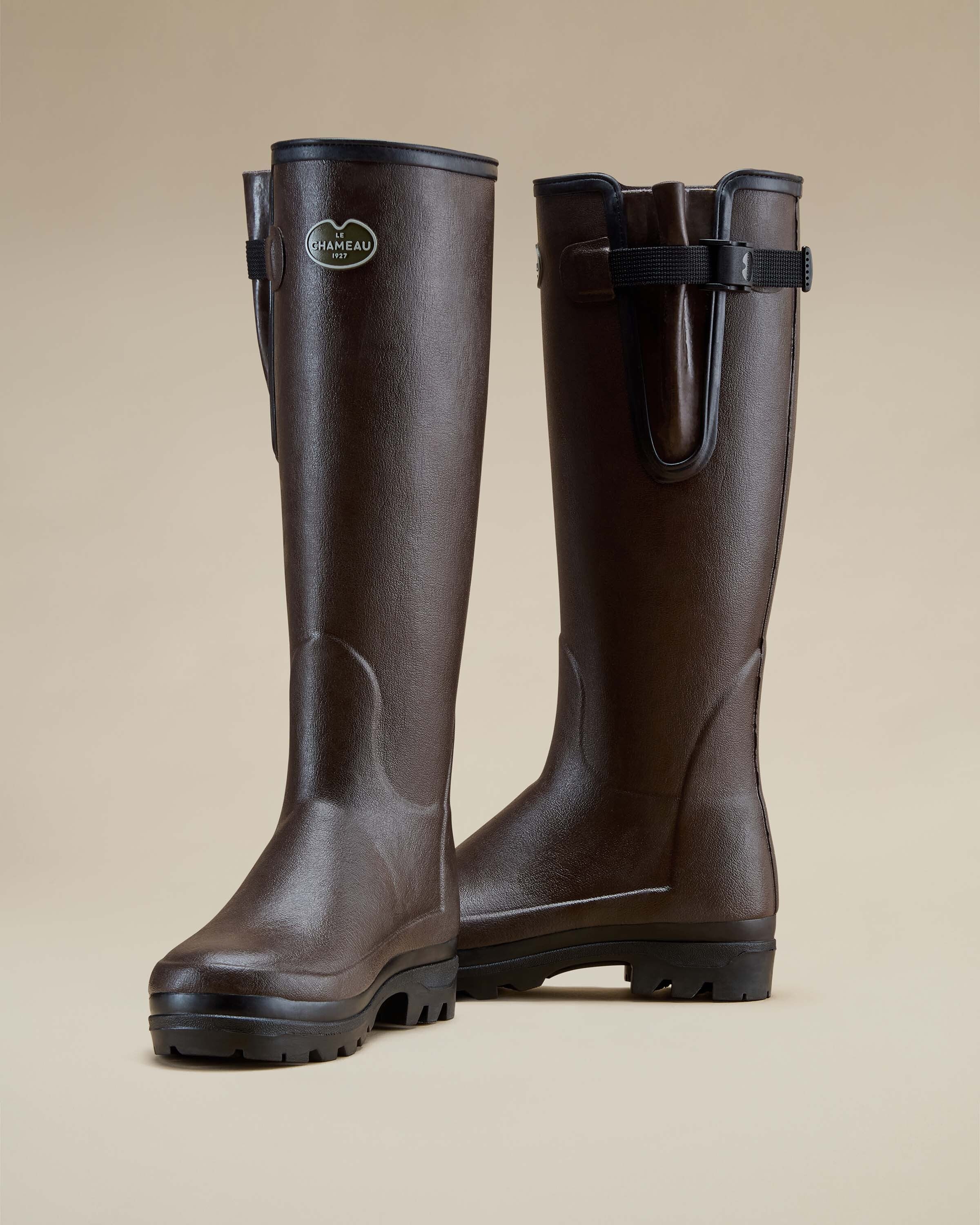 Two Women's Vierzonord Neoprene Lined Wellington Boot in Dark Brown posed upright in opposite orientations—one front-facing, the other with the gusset side visible—against a softly lit beige backdrop. This view accentuates the boots' symmetrical design, reinforced instep paneling, and practical details such as the adjustable side strap and the supportive arch structure of the midsole.